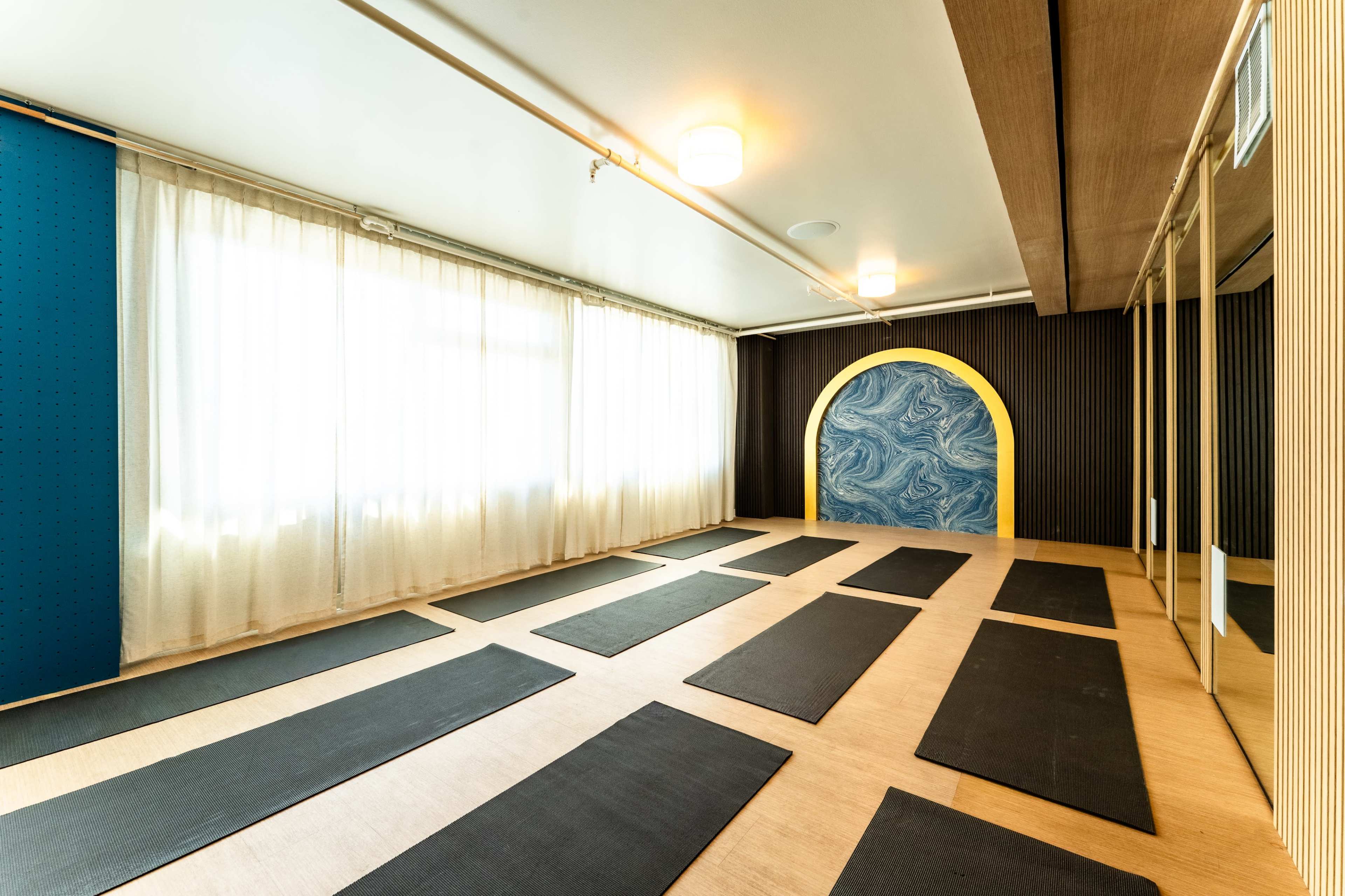 The image shows a yoga studio featuring wooden flooring, yoga mats arranged in parallel lines, large windows with sheer curtains, and a decorative archway with a blue and yellow design on the wall.