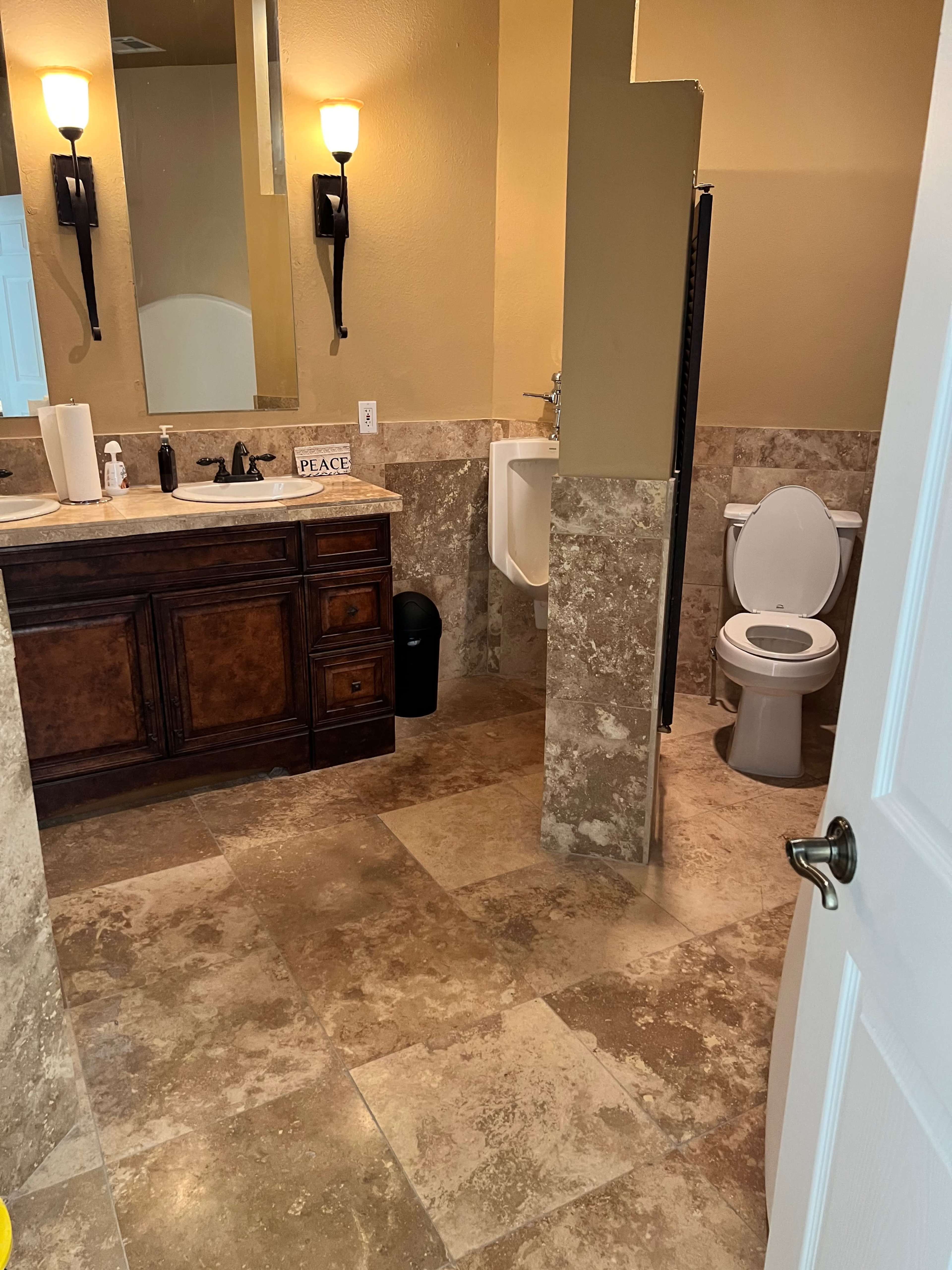 The image shows a bathroom with a toilet, a wall-mounted urinal, a sink with cabinets, and tiled flooring.