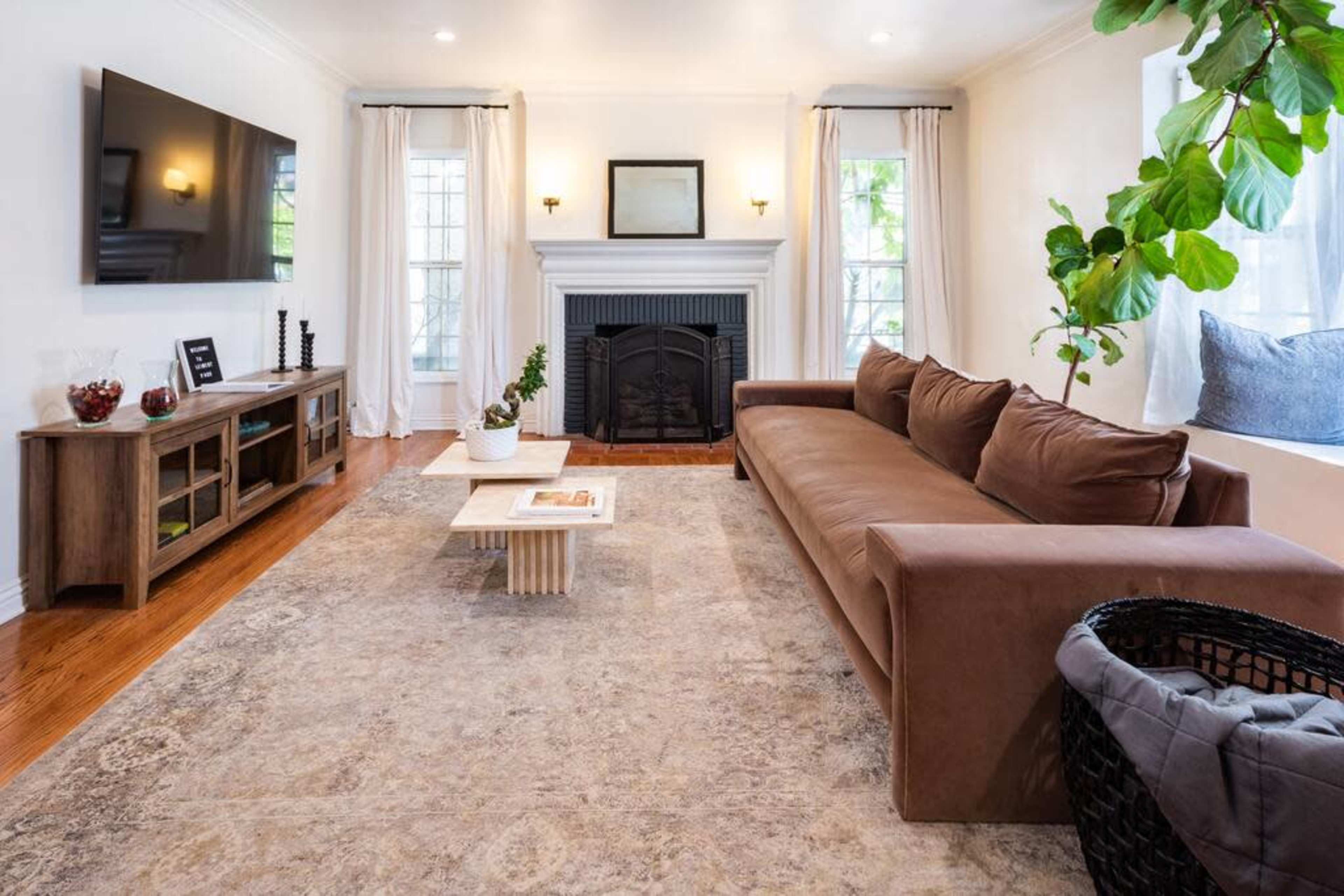The living room features a brown sofa facing a mounted television and a fireplace, with a coffee table and a large area rug on polished wooden floors.