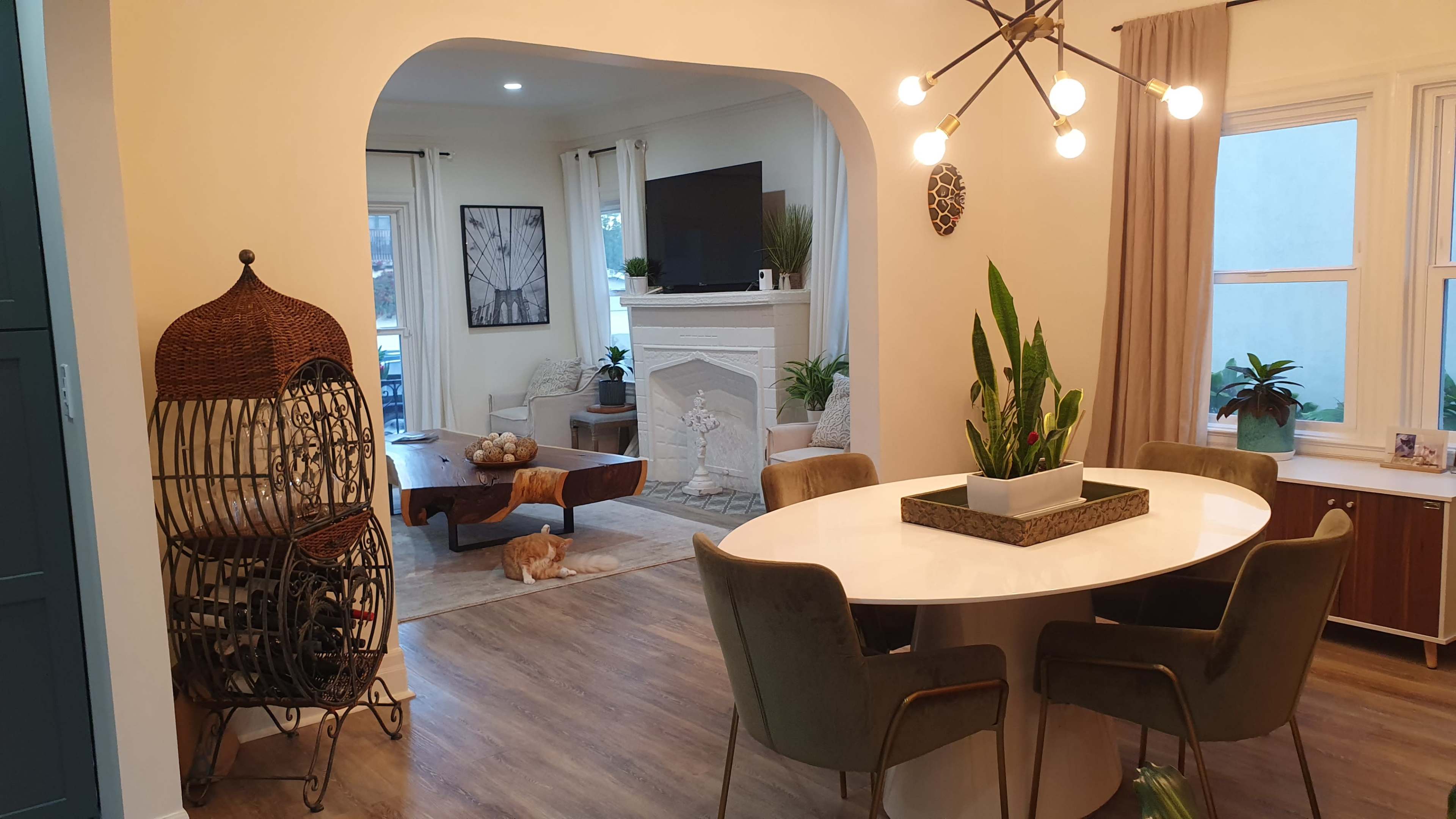 A modern dining area features a circular table surrounded by green chairs, with an archway leading to a living room that has a fireplace and plants.