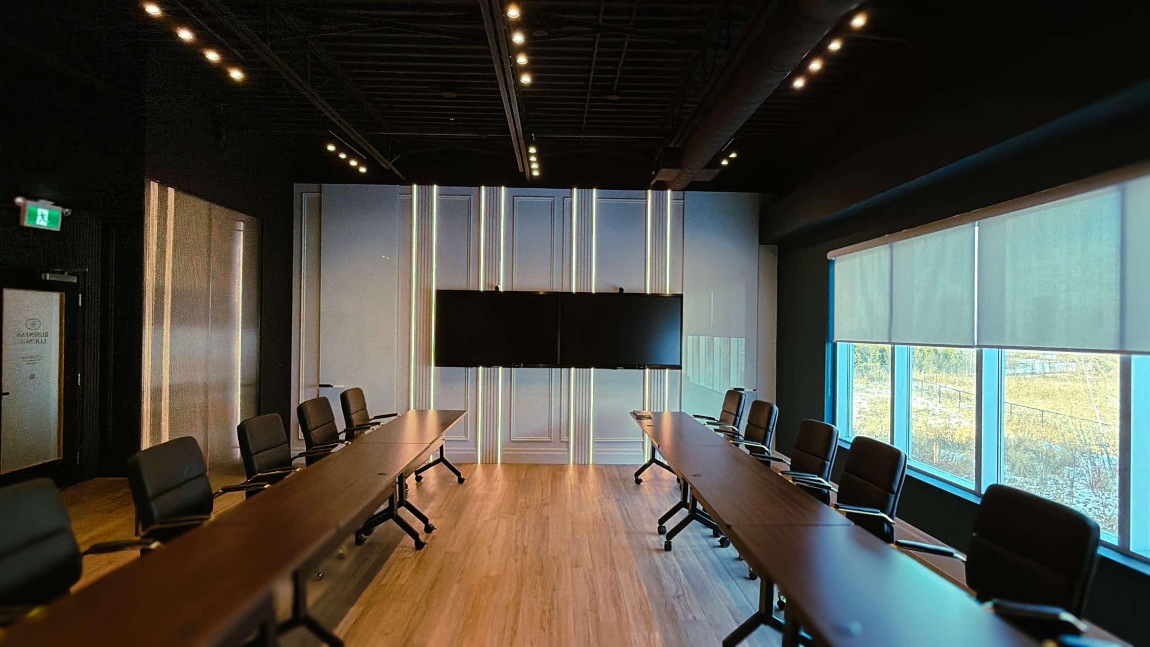 The image shows a modern conference room with a long table, black chairs, a large screen on the wall, and windows letting in natural light.