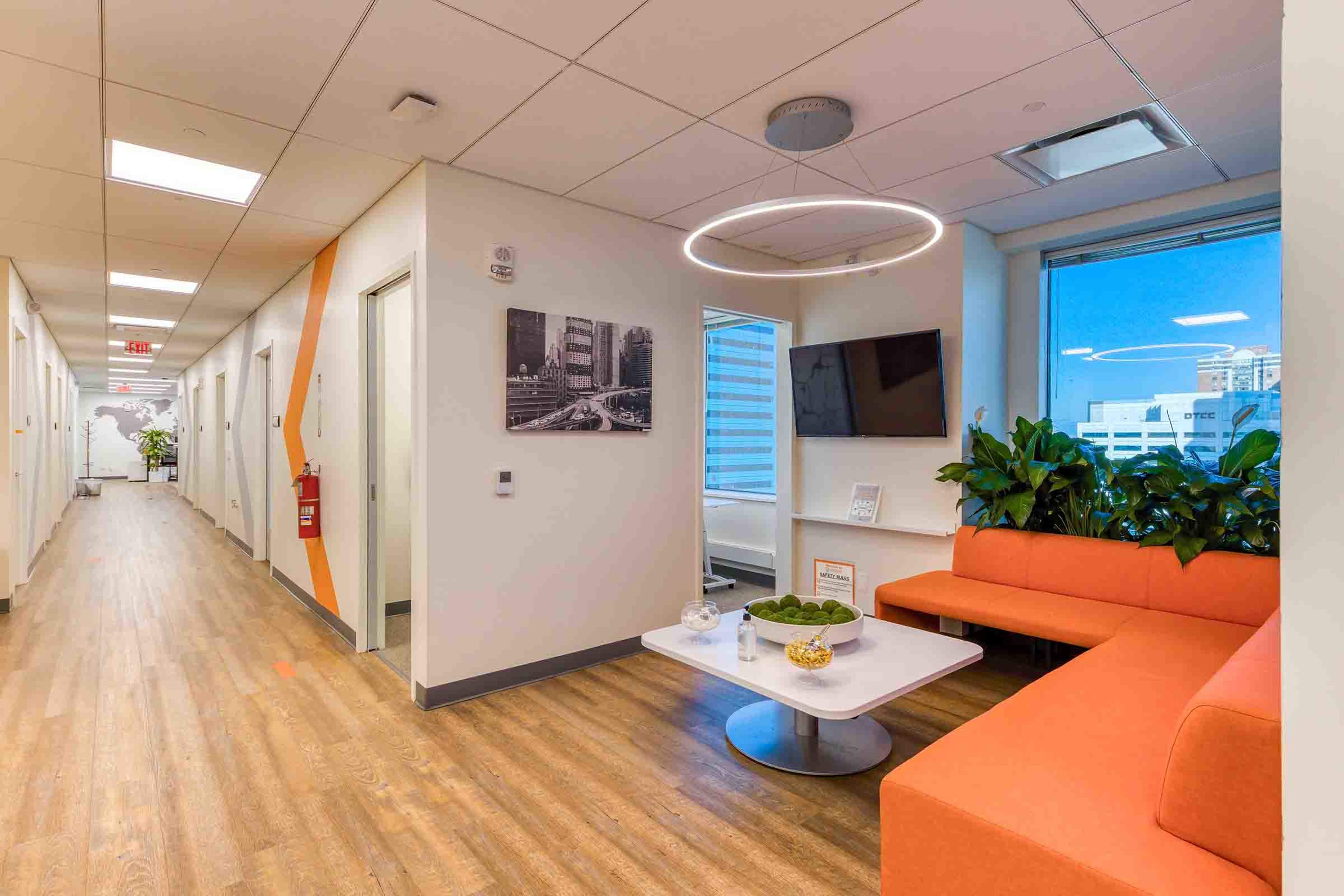 A modern office hallway with orange seating, a wall-mounted TV, and large windows providing natural light.