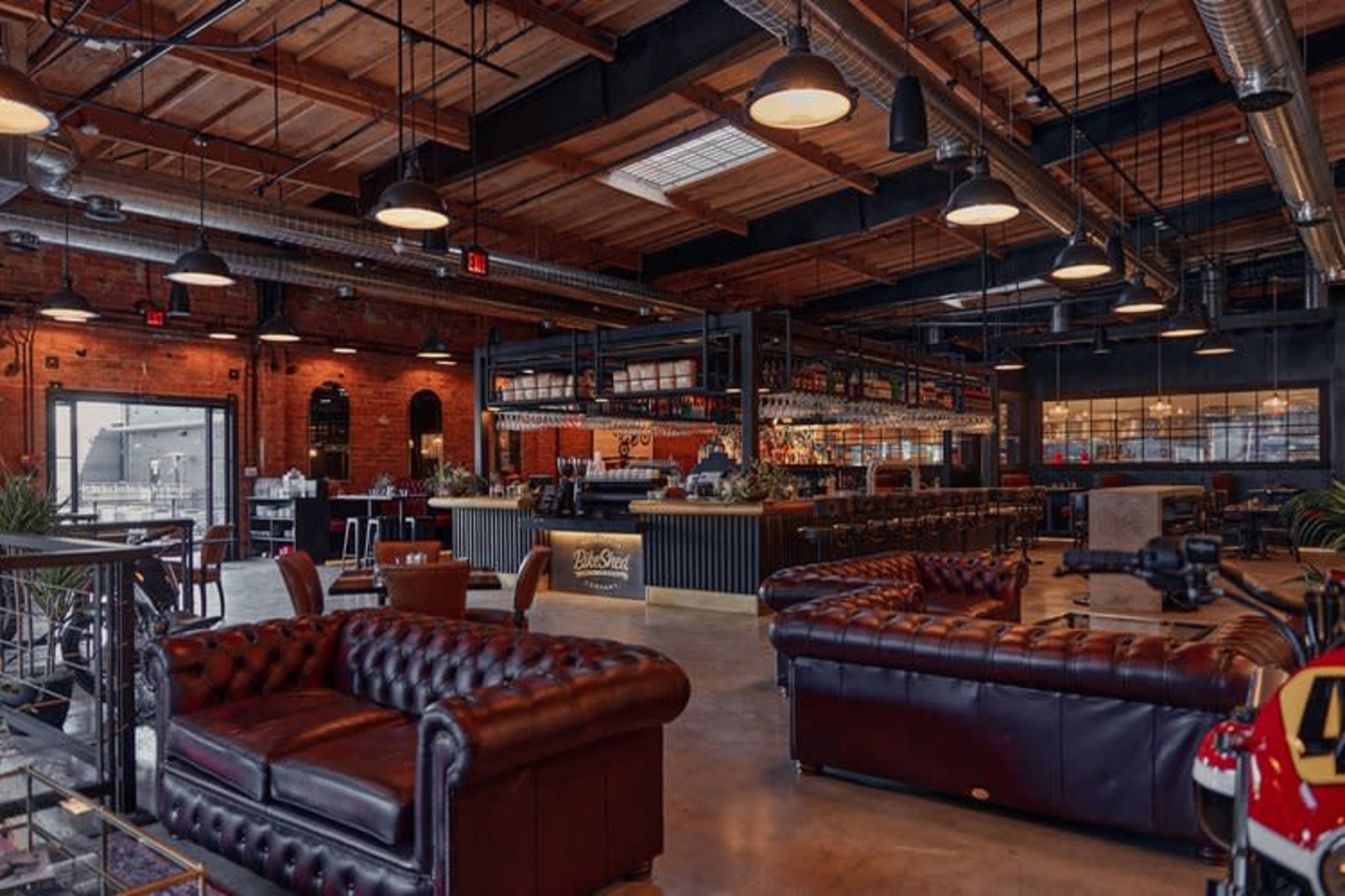 The image shows a modern bar interior featuring leather couches, a long bar with shelves of liquor, and an industrial-style design with exposed beams and large windows.