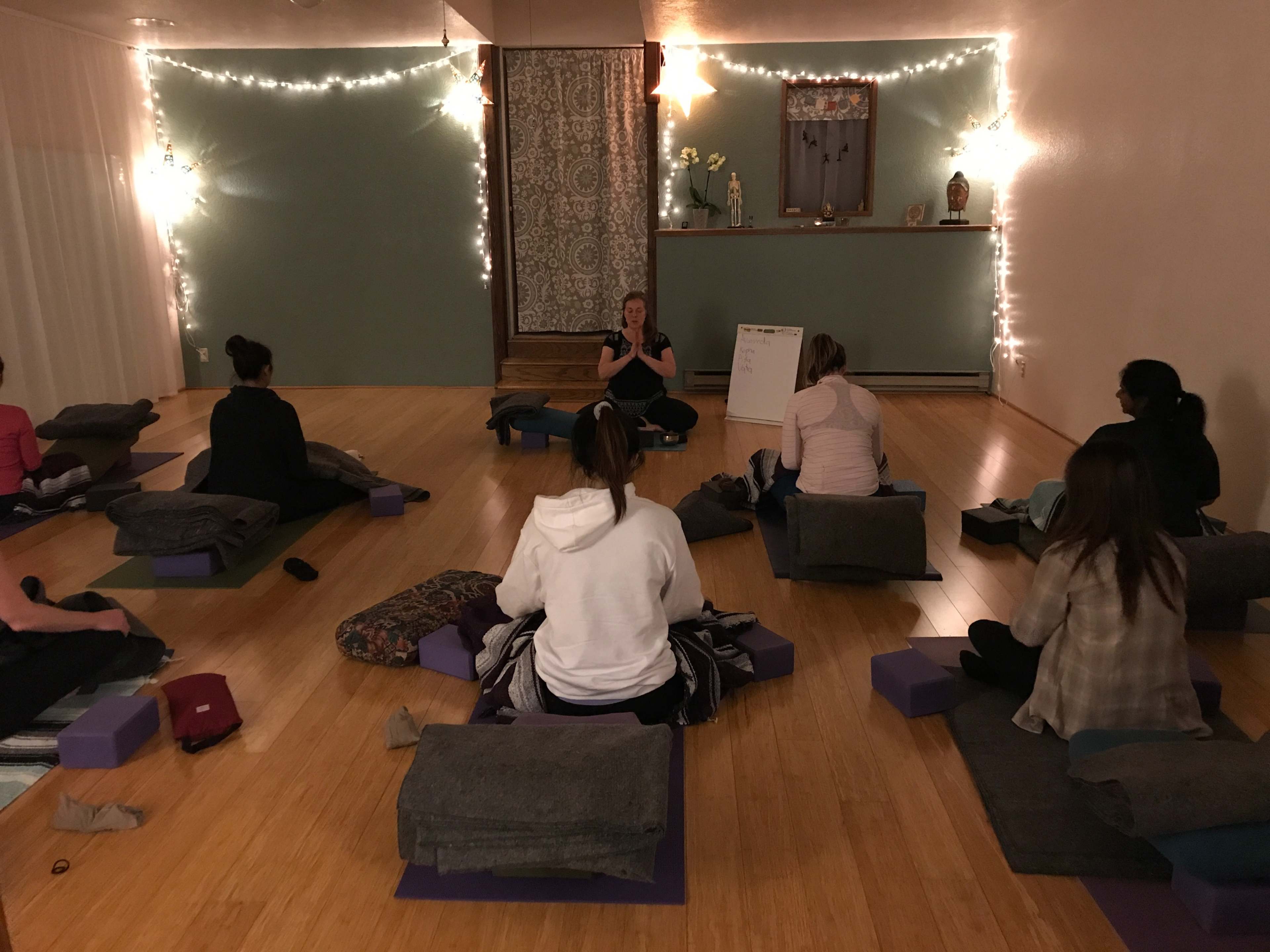 A yoga instructor leads a class in a softly lit studio, with participants seated on mats and blankets.