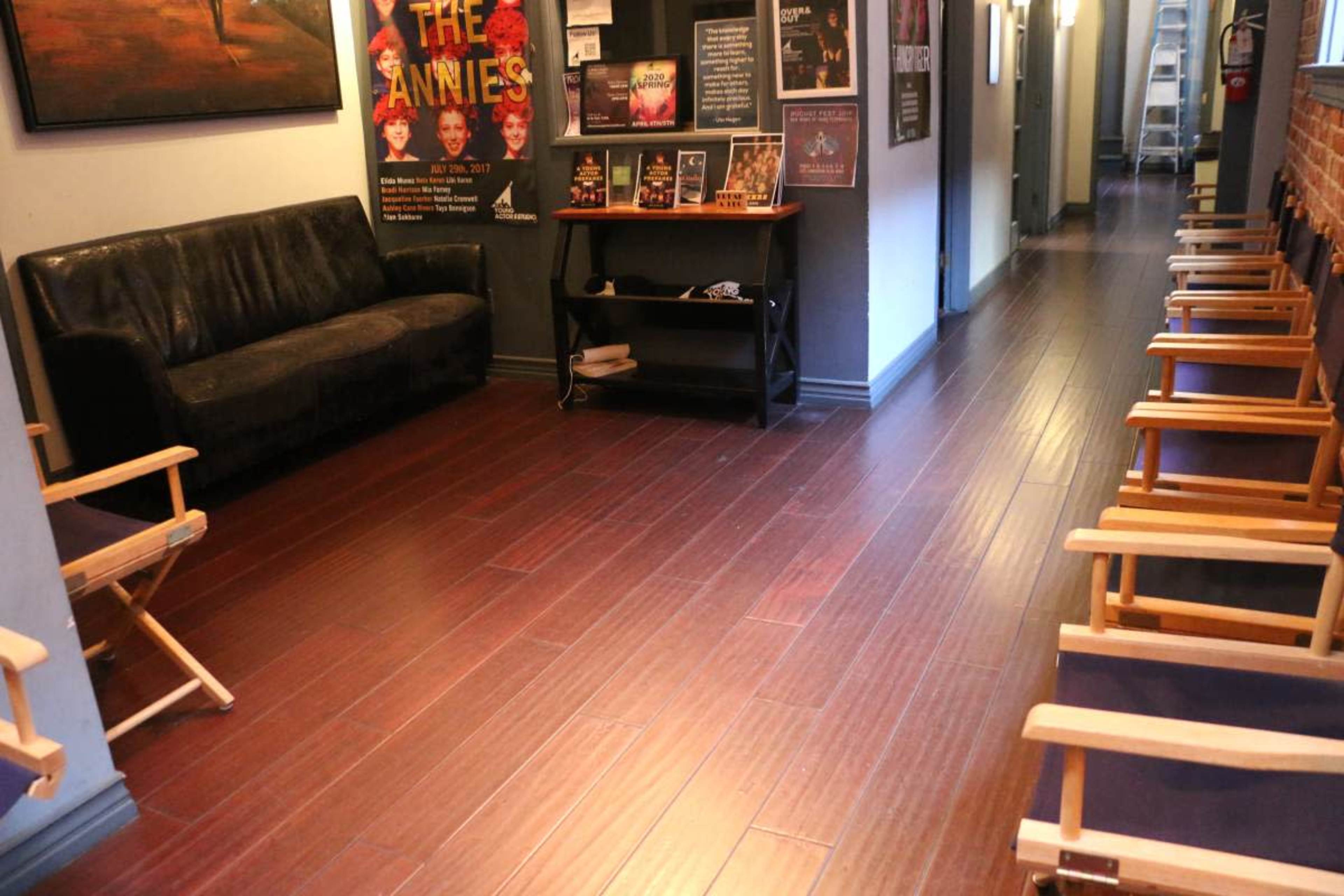 The image shows a hallway with a dark couch, a wooden table, and several empty director's chairs lined along the side.