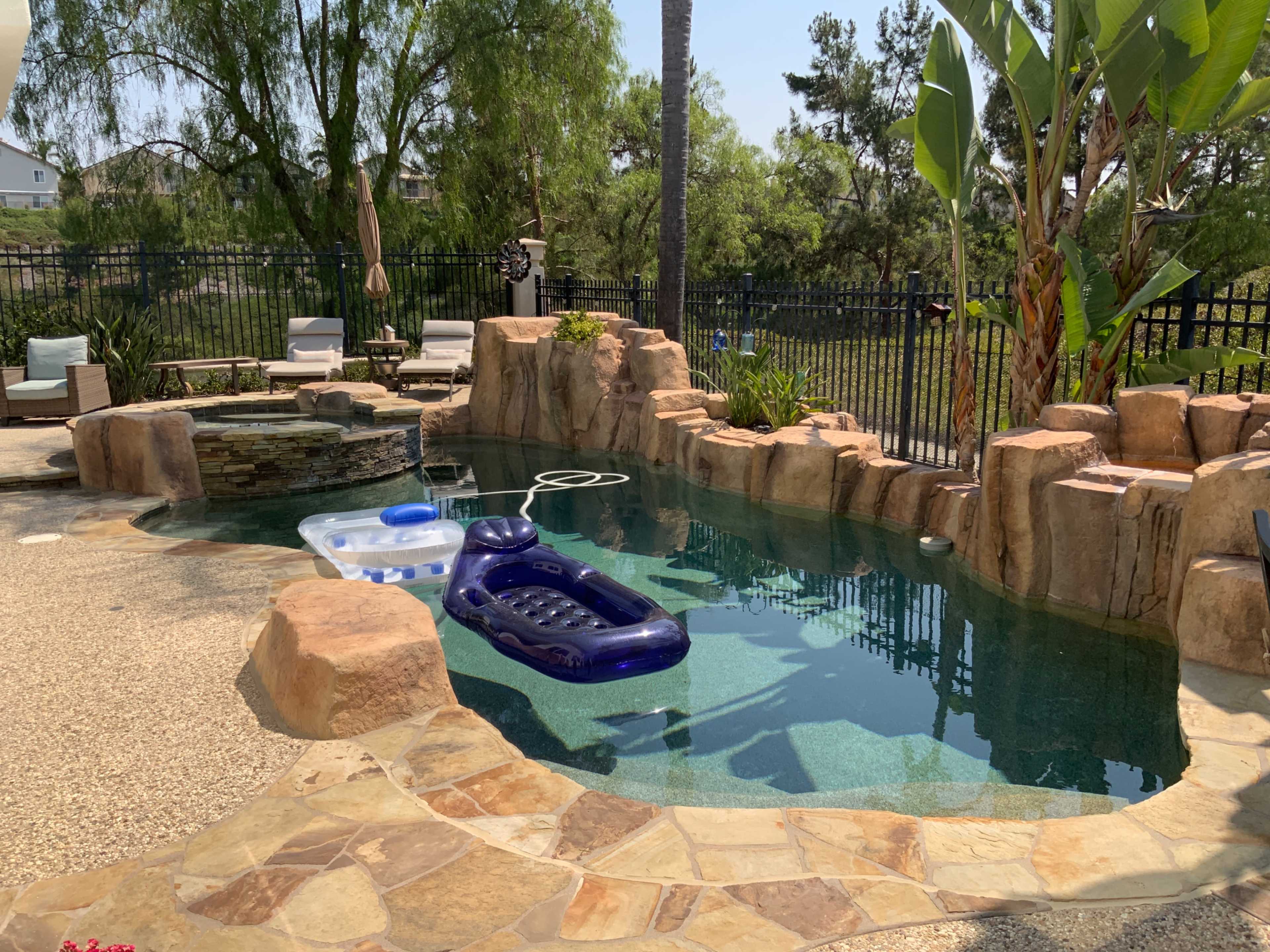 The image shows a backyard pool surrounded by rock formations, lounge chairs, and tropical plants.