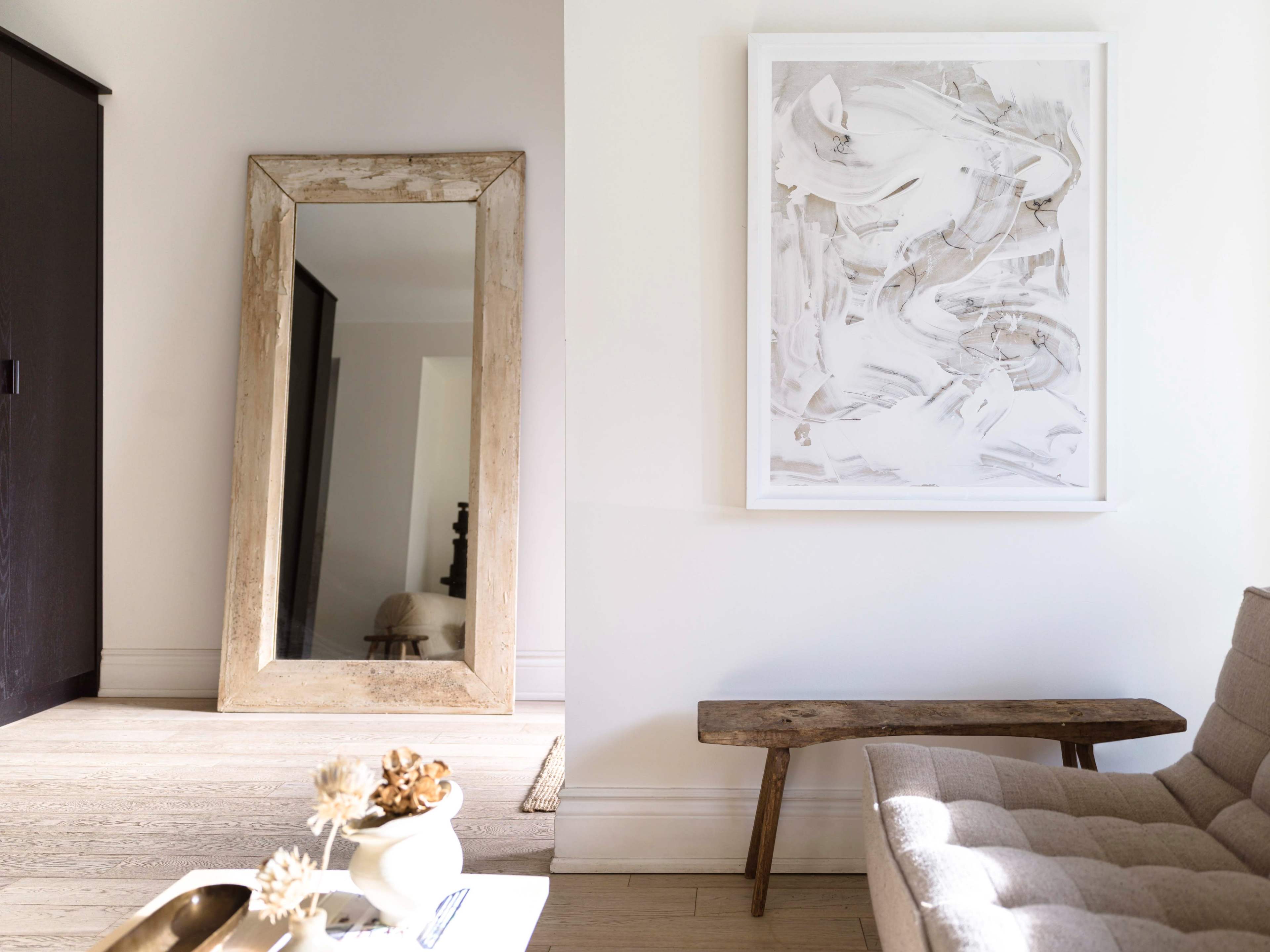 A large rustic mirror stands against the wall next to a simple wooden bench and a piece of abstract artwork hanging above it.
