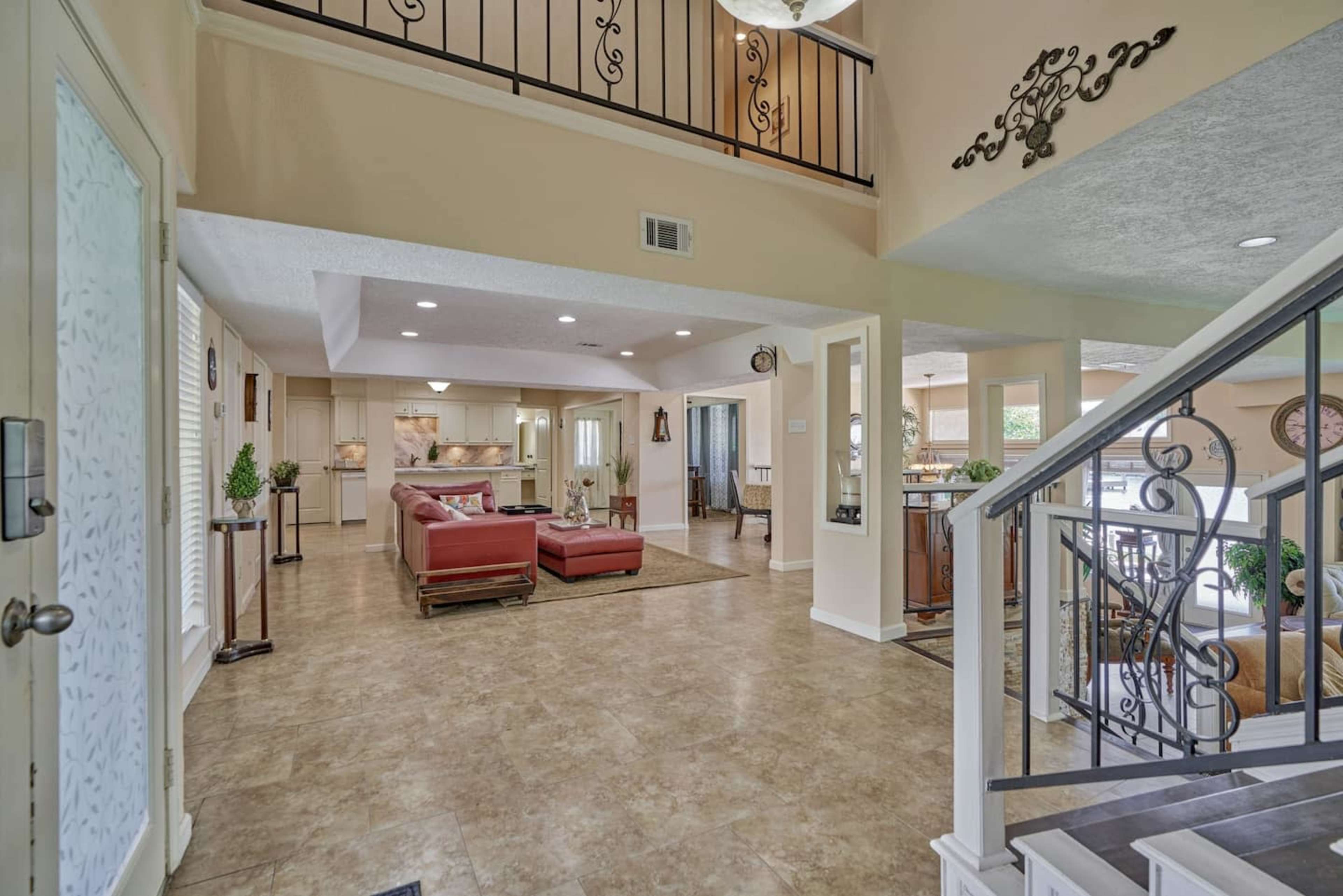 The image shows a spacious, open foyer with a staircase leading to an upper level, framed by a living area featuring a red sofa and brown furnishings.