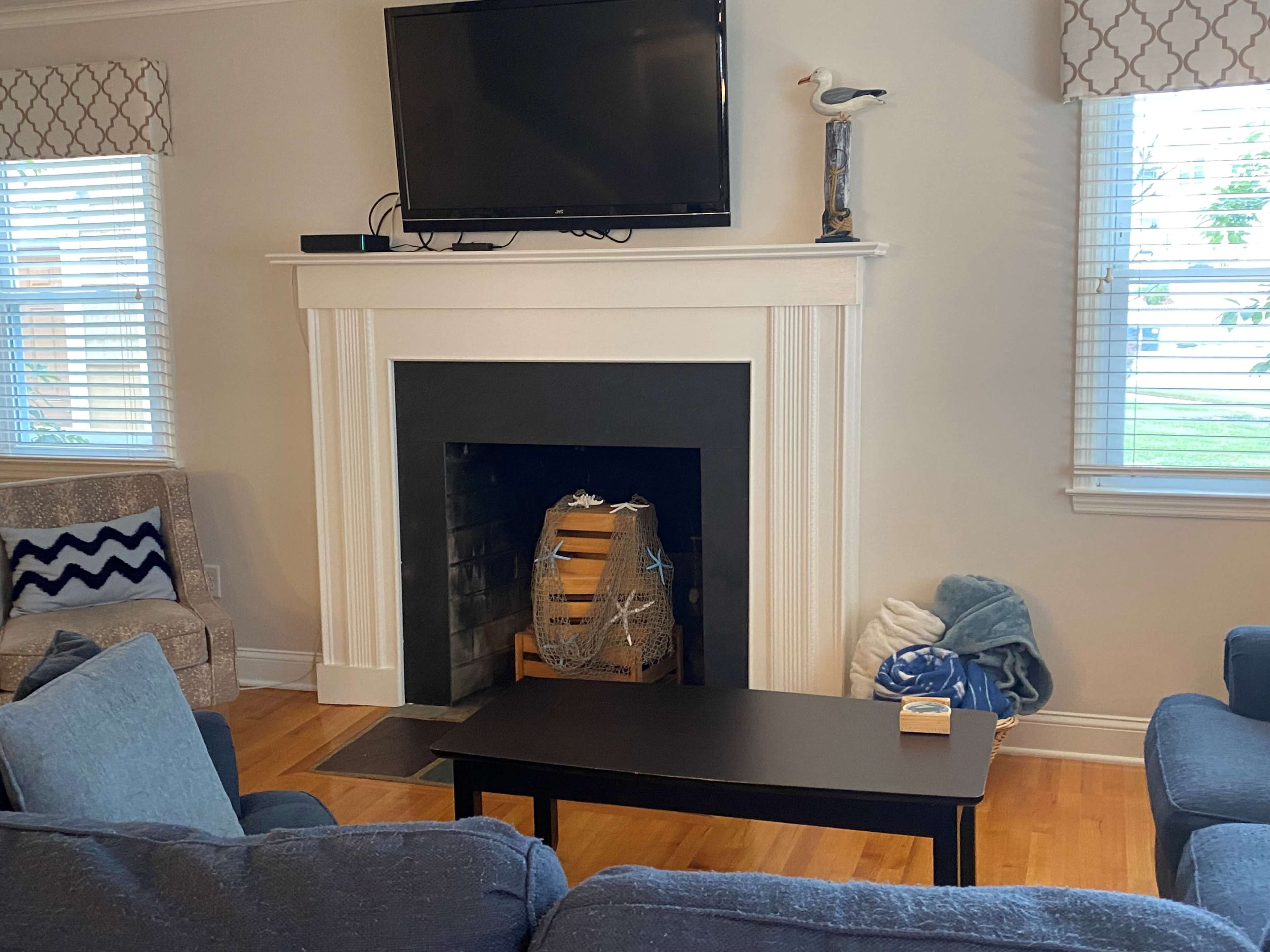 The living room features a fireplace with a wooden crate inside, a black coffee table, and a mounted television above the mantel, with light streaming through the windows.