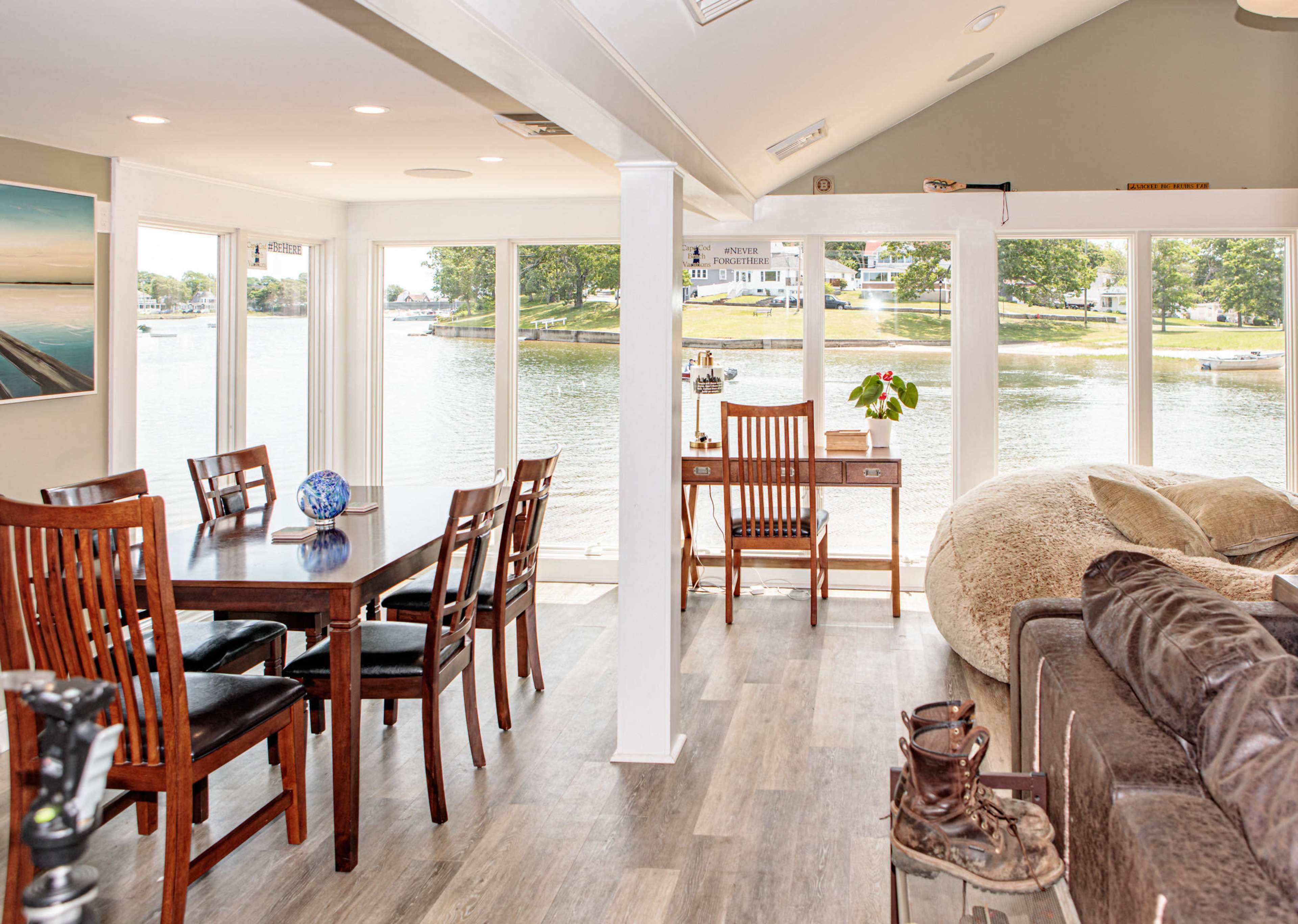 The interior of a bright, open-concept living space features a dining area, a desk by large windows overlooking a lake, and a cozy seating area with a couch.