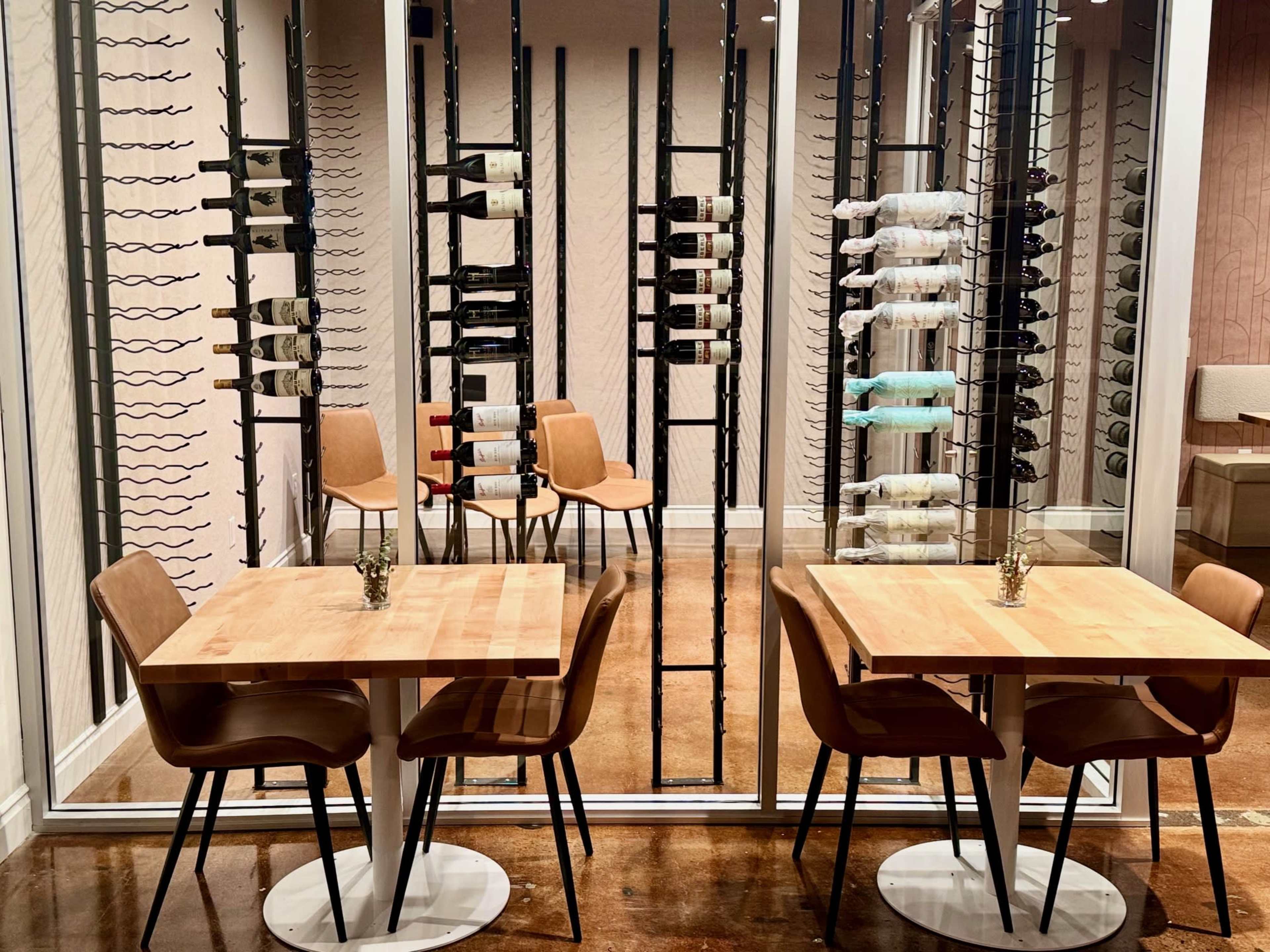 The image shows a modern dining area with two wooden tables and chairs, framed by a wine rack displaying various bottles.