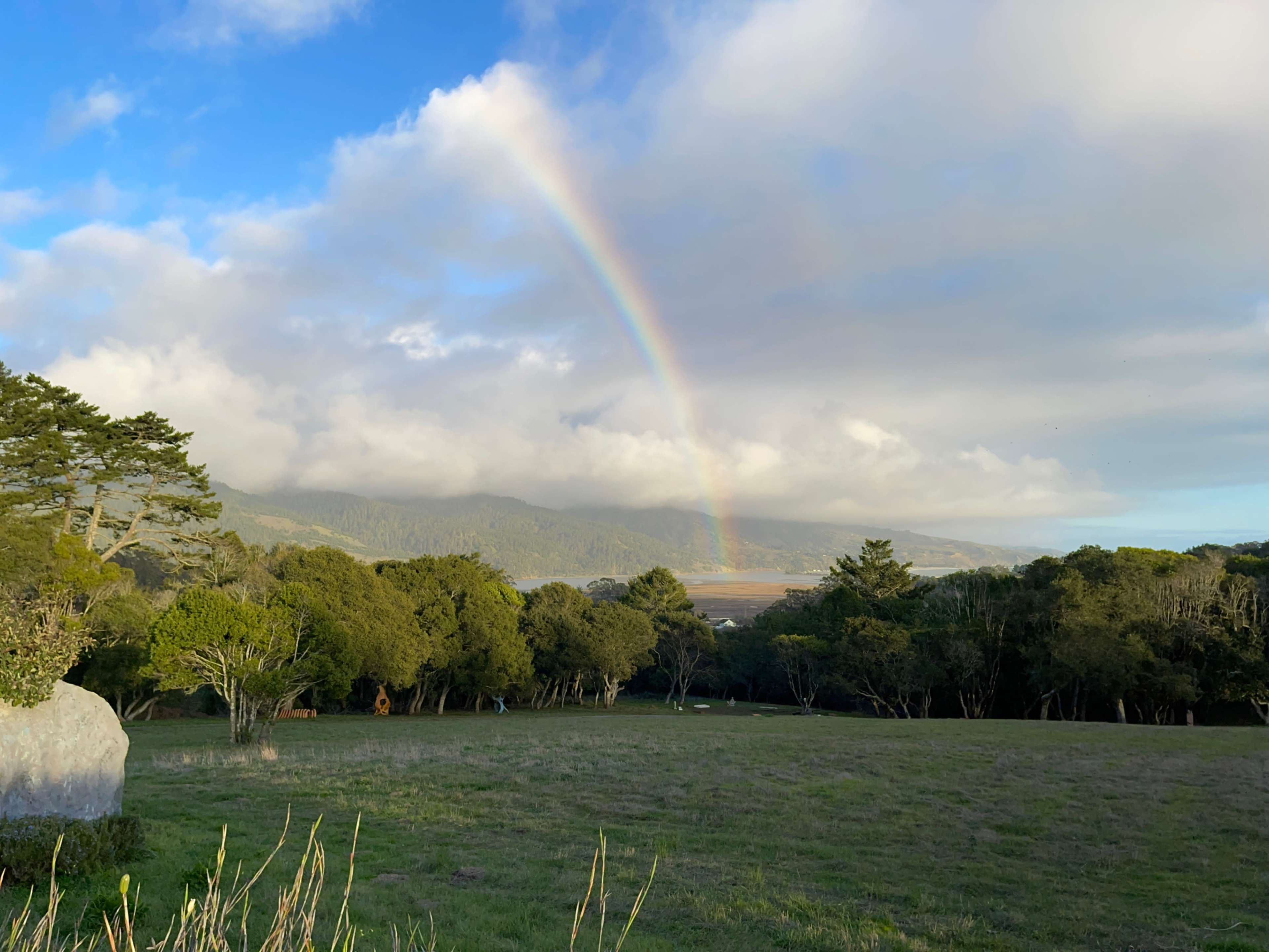 A rainbow arches over a green landscape with trees and hills under a partly cloudy sky.