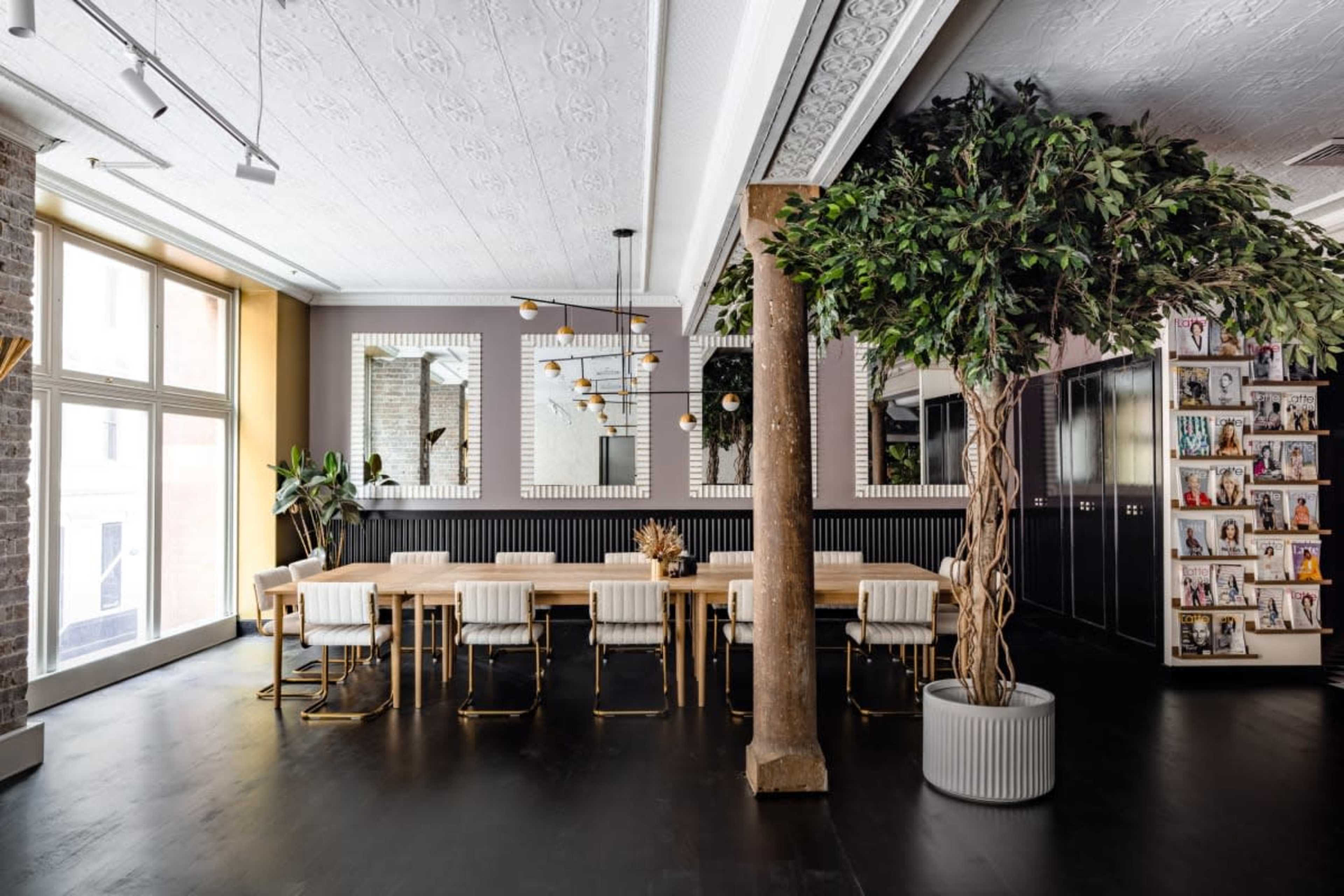 A modern interior featuring a long wooden table surrounded by white chairs, with large windows, decorative plants, and shelves filled with magazines.