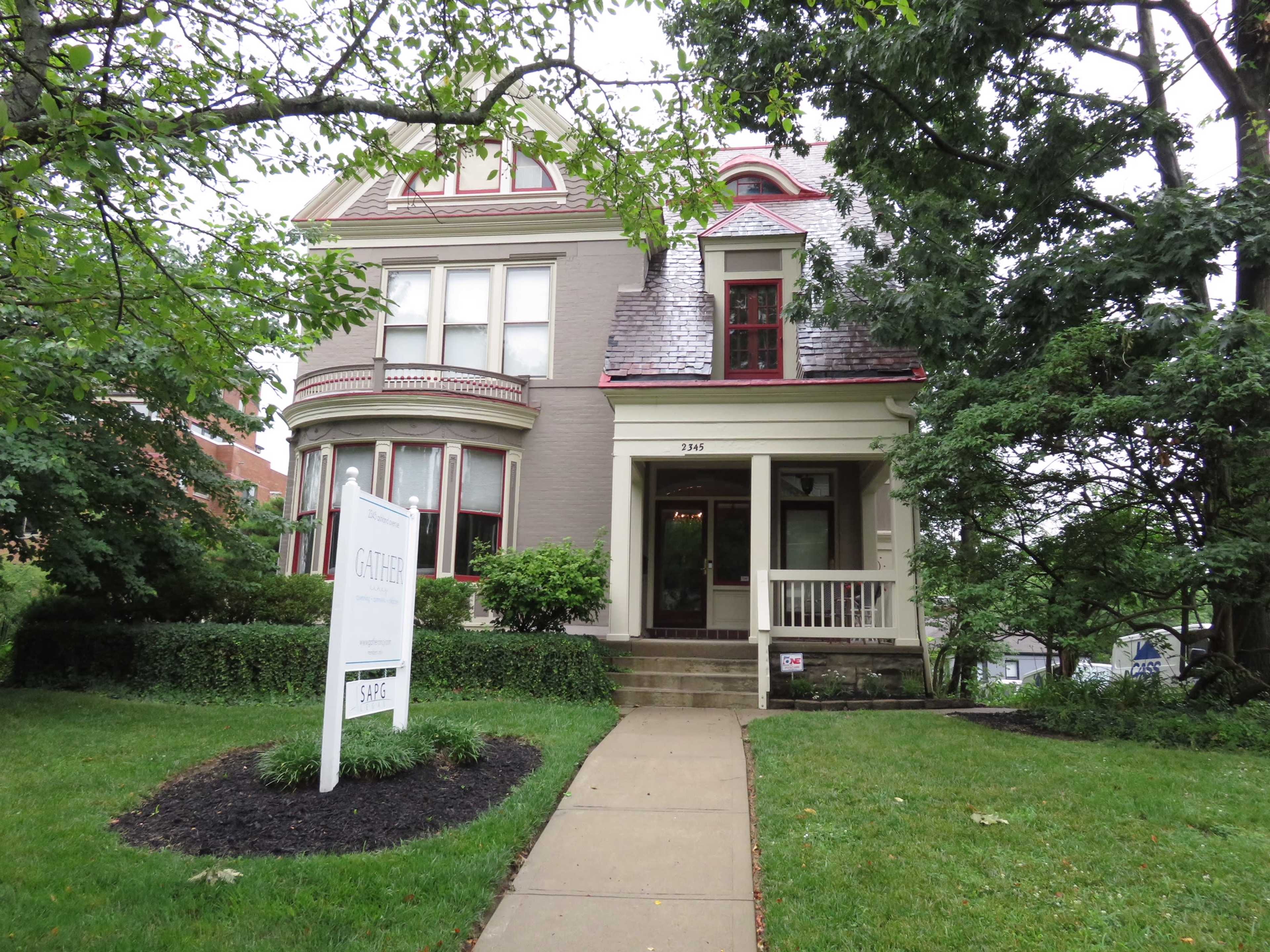 A large Victorian-style house with a peaked roof and red accents is surrounded by greenery, featuring a sign in front indicating it is for sale.