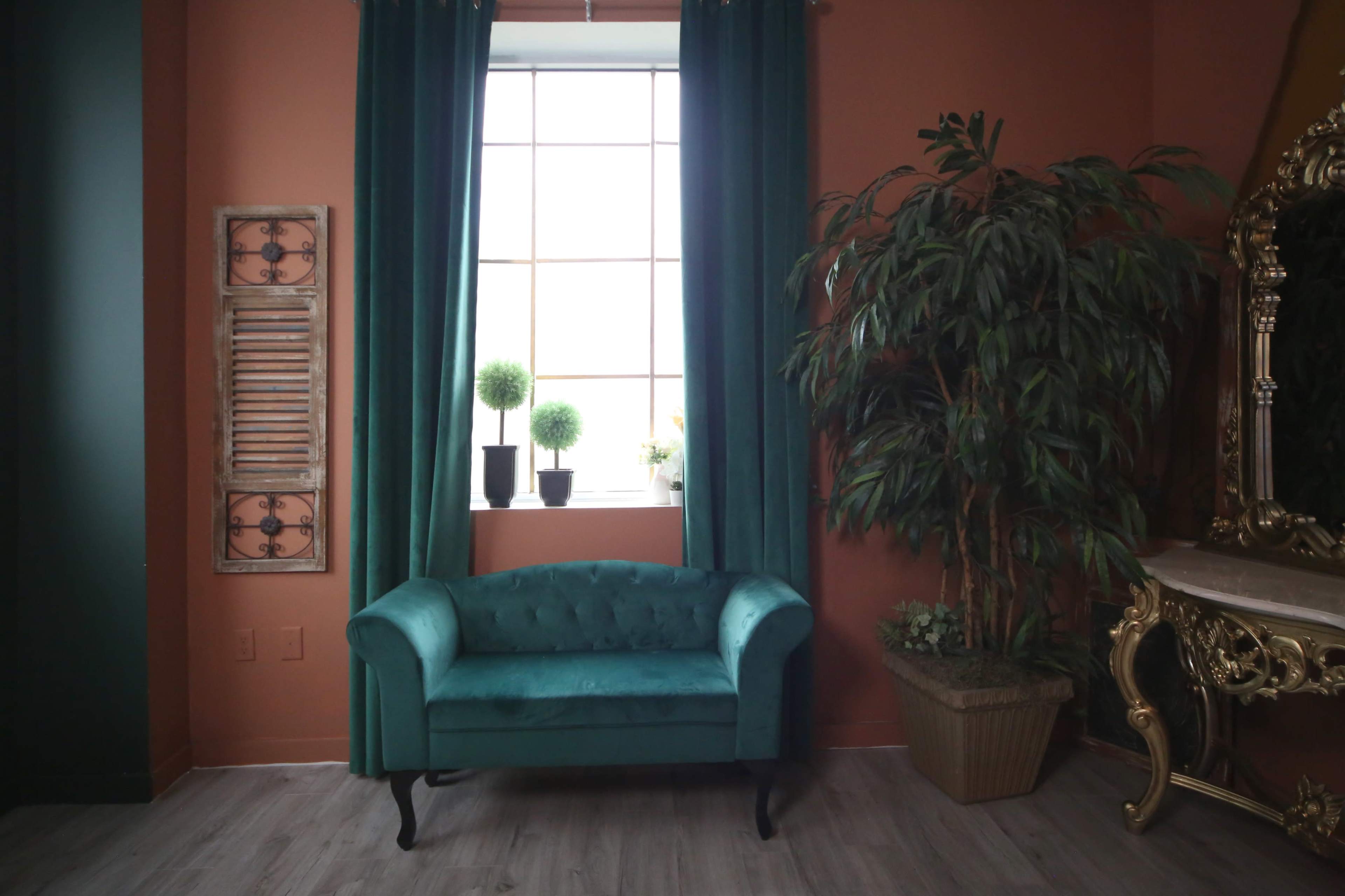 A turquoise loveseat sits in front of a large window, flanked by green curtains and potted plants.