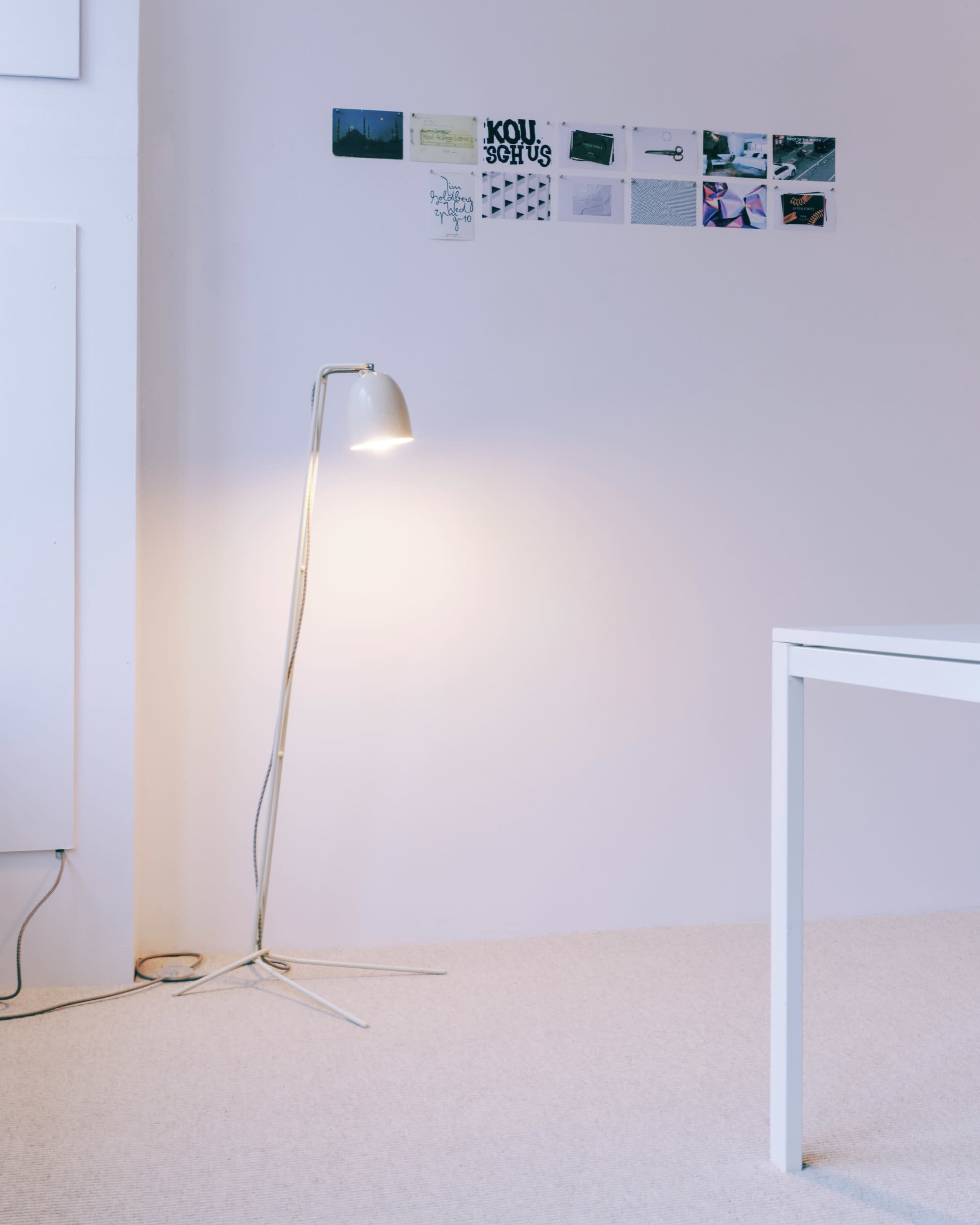 A floor lamp stands in the corner of a room next to a wall covered with a series of postcards and images, while a white table is positioned nearby.