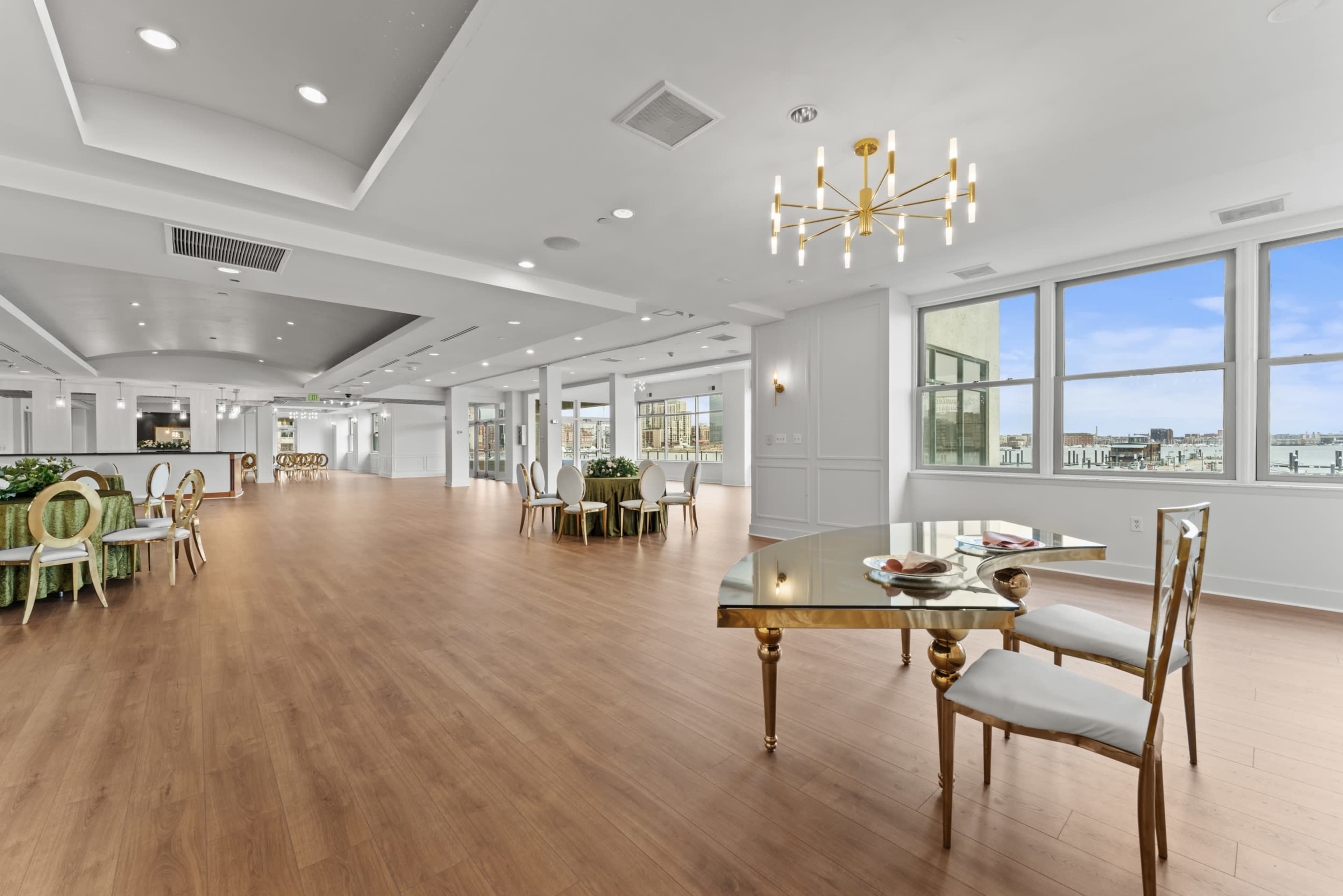 The image shows a spacious interior room featuring wooden flooring, large windows, and elegant furniture, including a gold-accented table and stylish chairs.