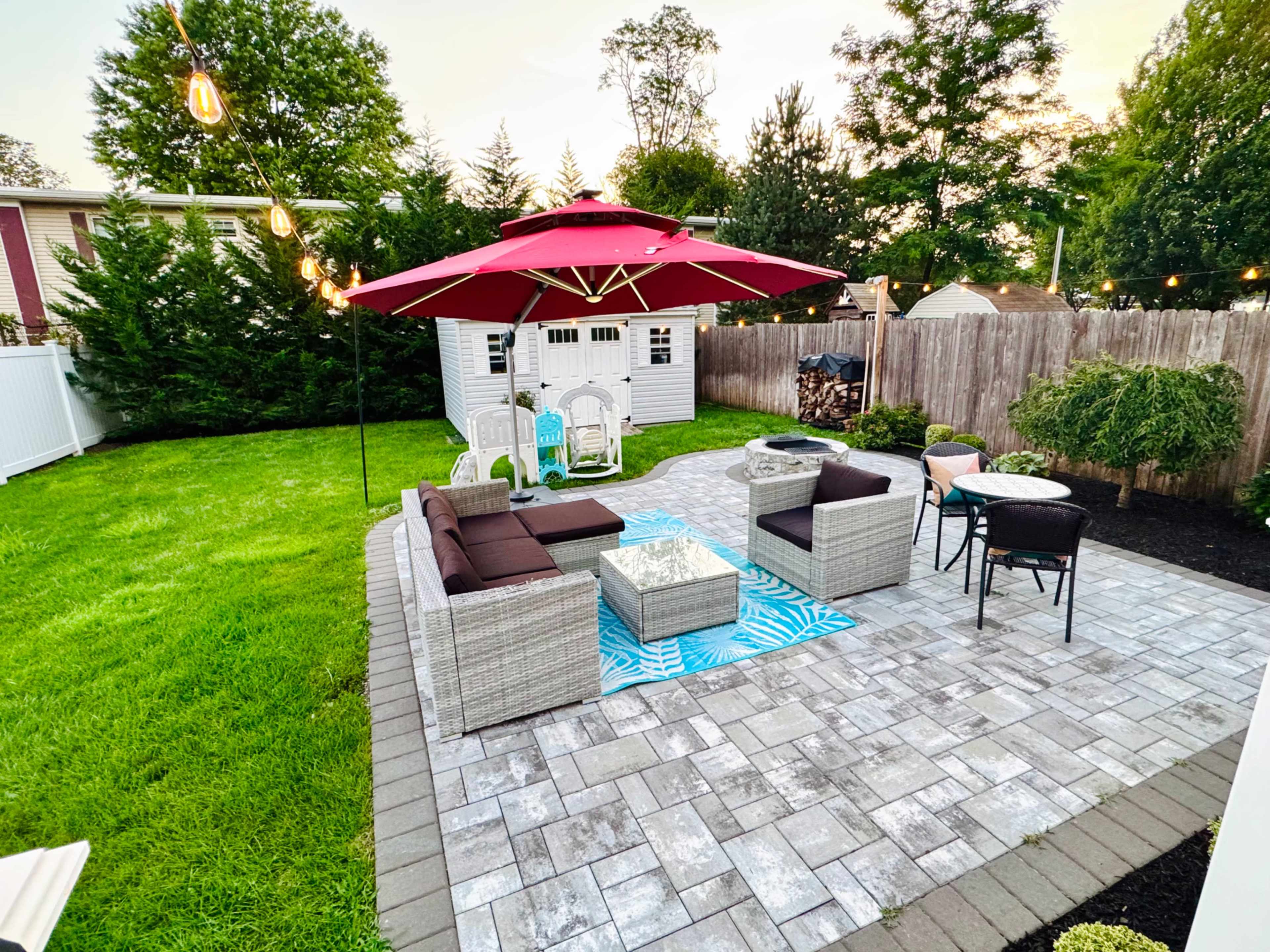 A backyard patio features a seating area with modern furniture, an umbrella, a small table, and a nearby shed, surrounded by green grass and hedges.