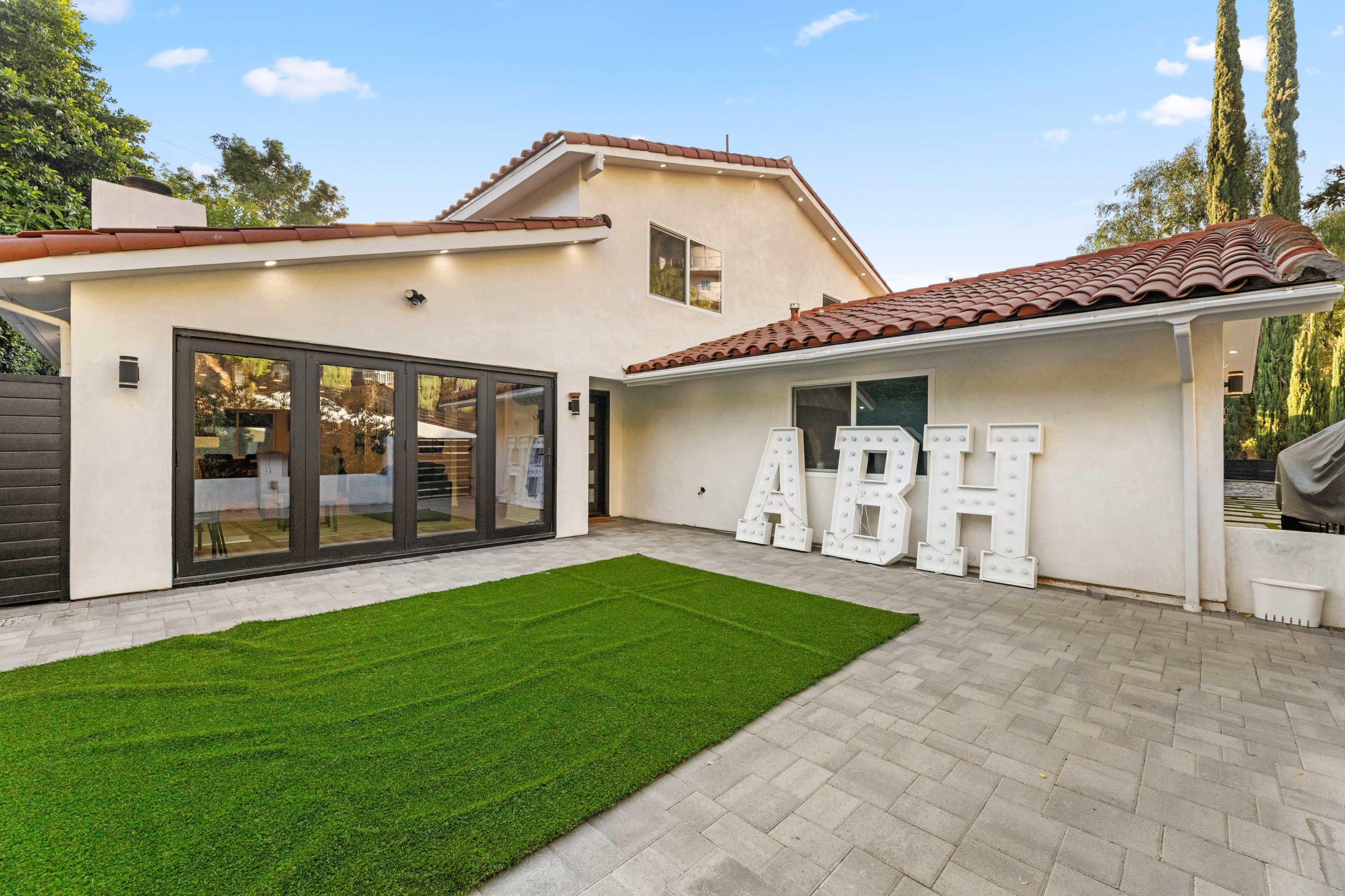 The image shows a modern outdoor patio area with a green artificial lawn, a white wall featuring large, backlit letters spelling "ABH," and a house with a tiled roof in the background.