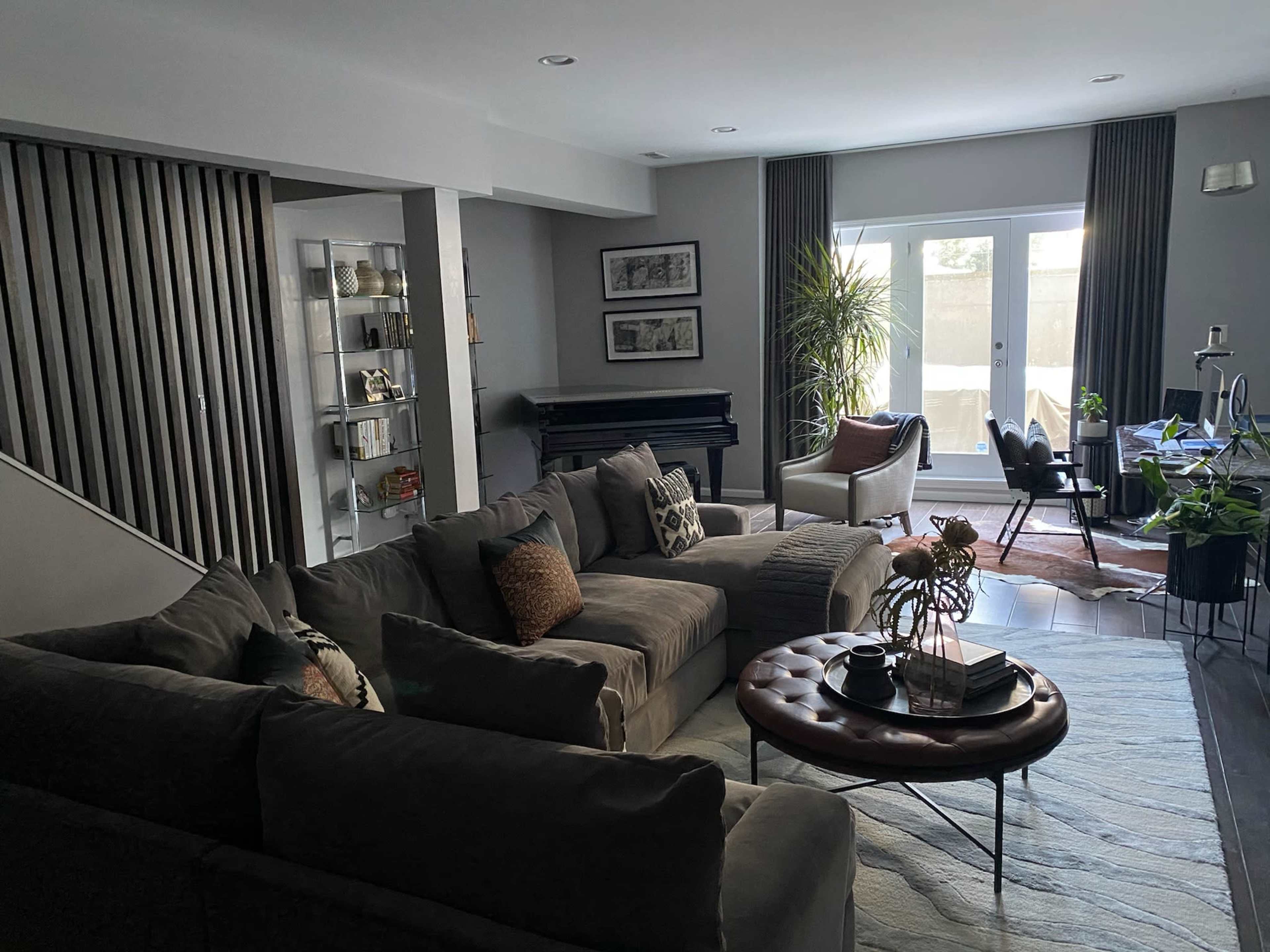 A modern living room featuring a large sectional sofa, a piano, and glass doors leading to a patio, with plants and decor throughout the space.