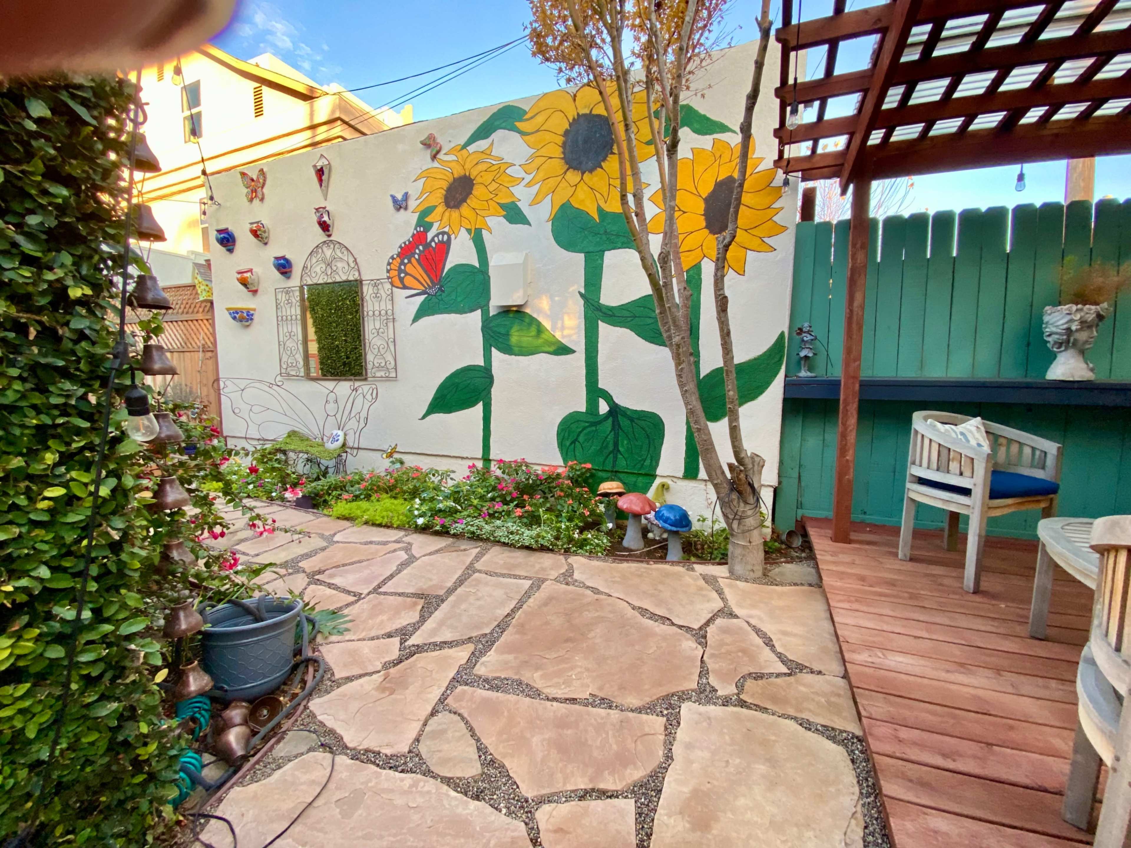 The image depicts a garden area with a stone pathway, colorful flowers, a mural of sunflowers, and a wooden deck with seating.