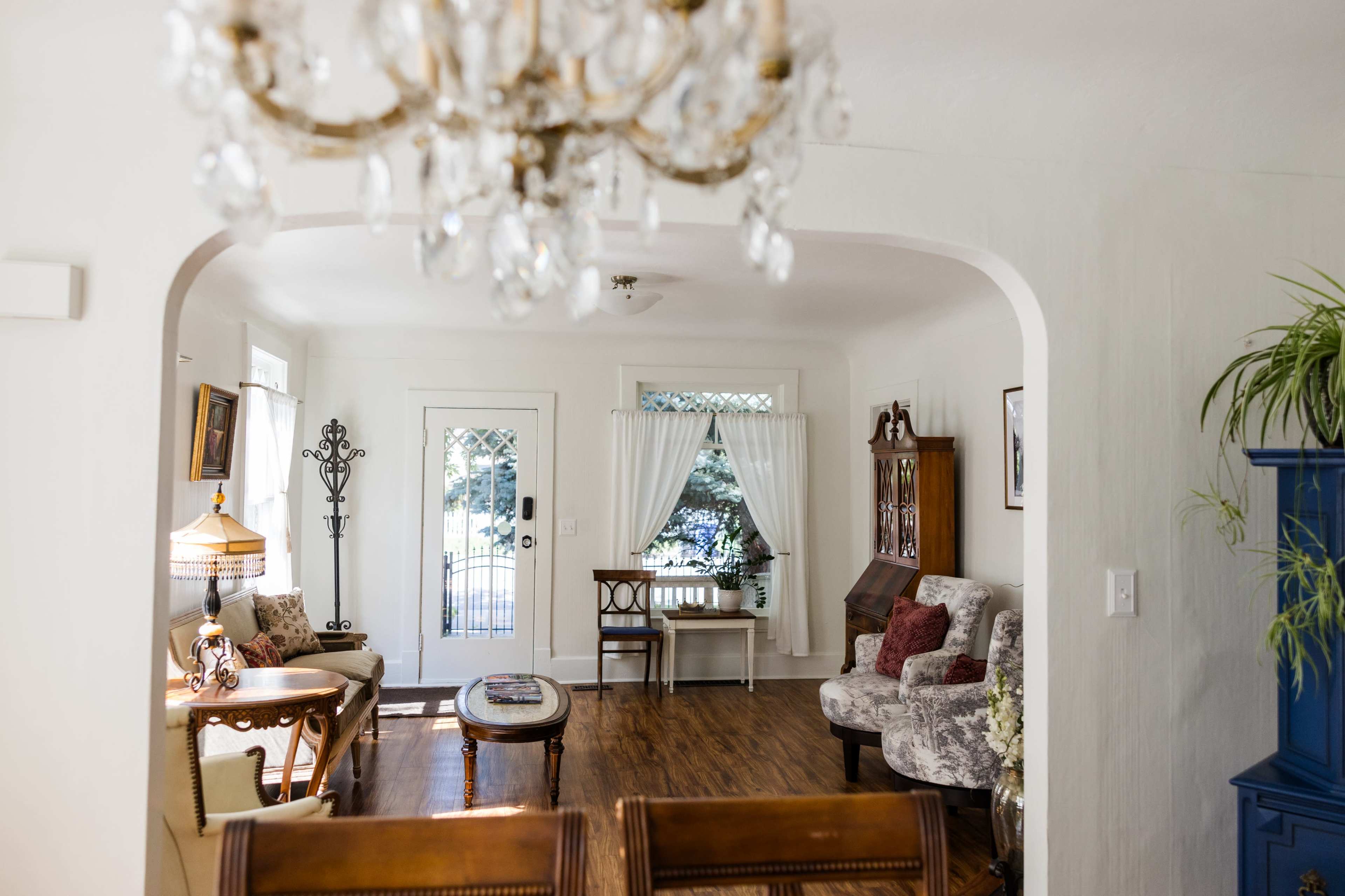 A bright living room with a chandelier, a wooden table, and furniture arranged around large windows facing a door that leads outside.