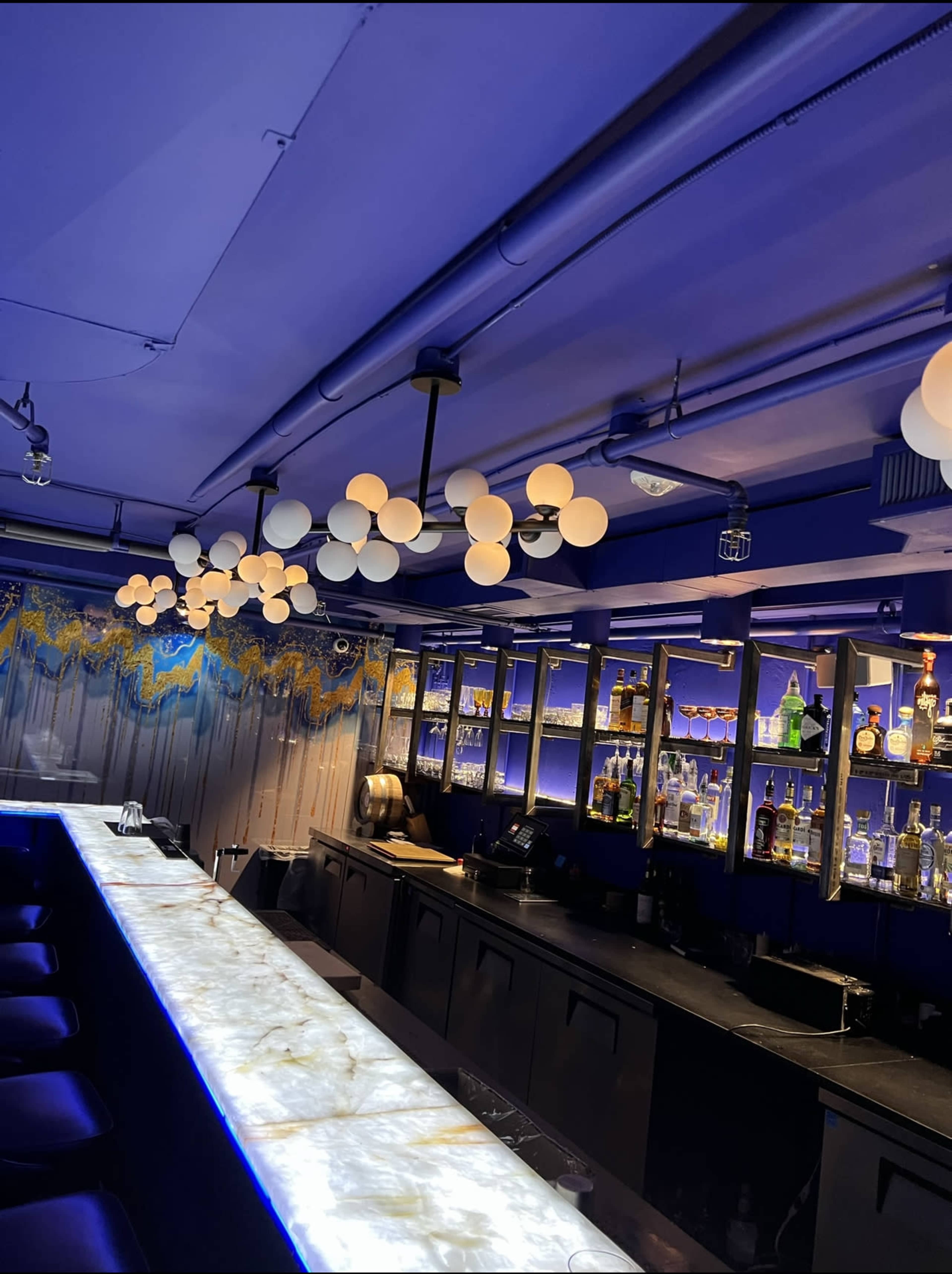 The image shows a modern bar with a marble countertop, illuminated by round pendant lights against a backdrop of blue walls and shelves stocked with various bottles.
