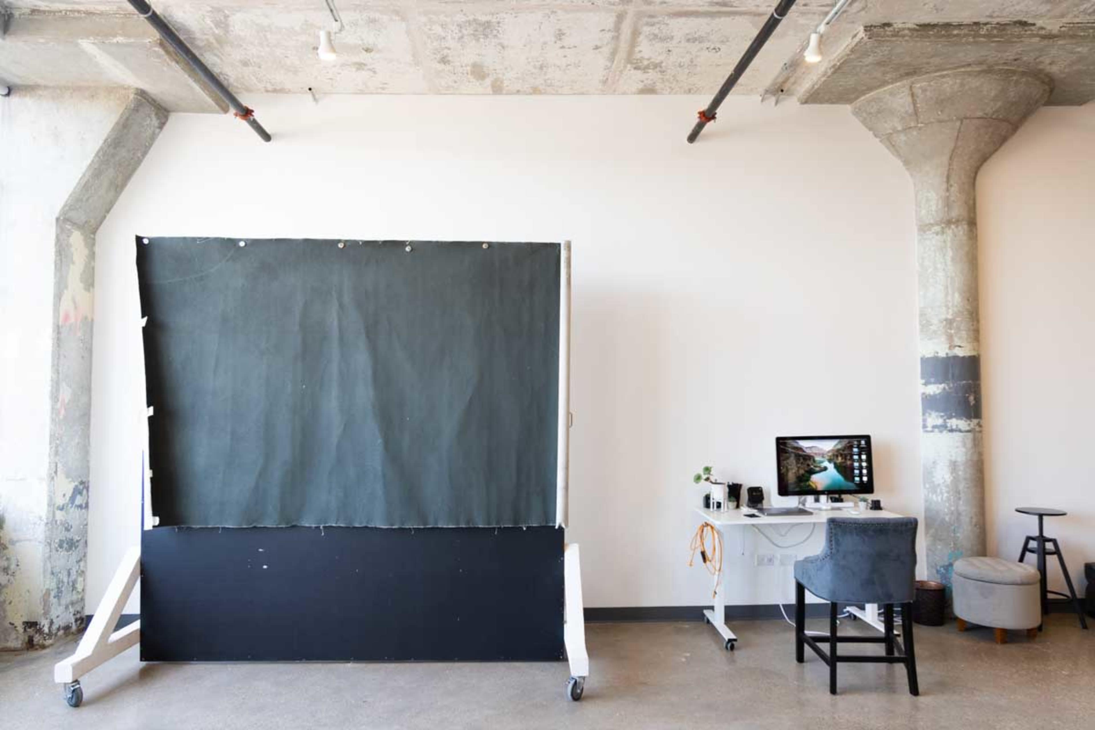 A studio space features a large, dark backdrop on wheels and a desk with a computer against a plain wall.