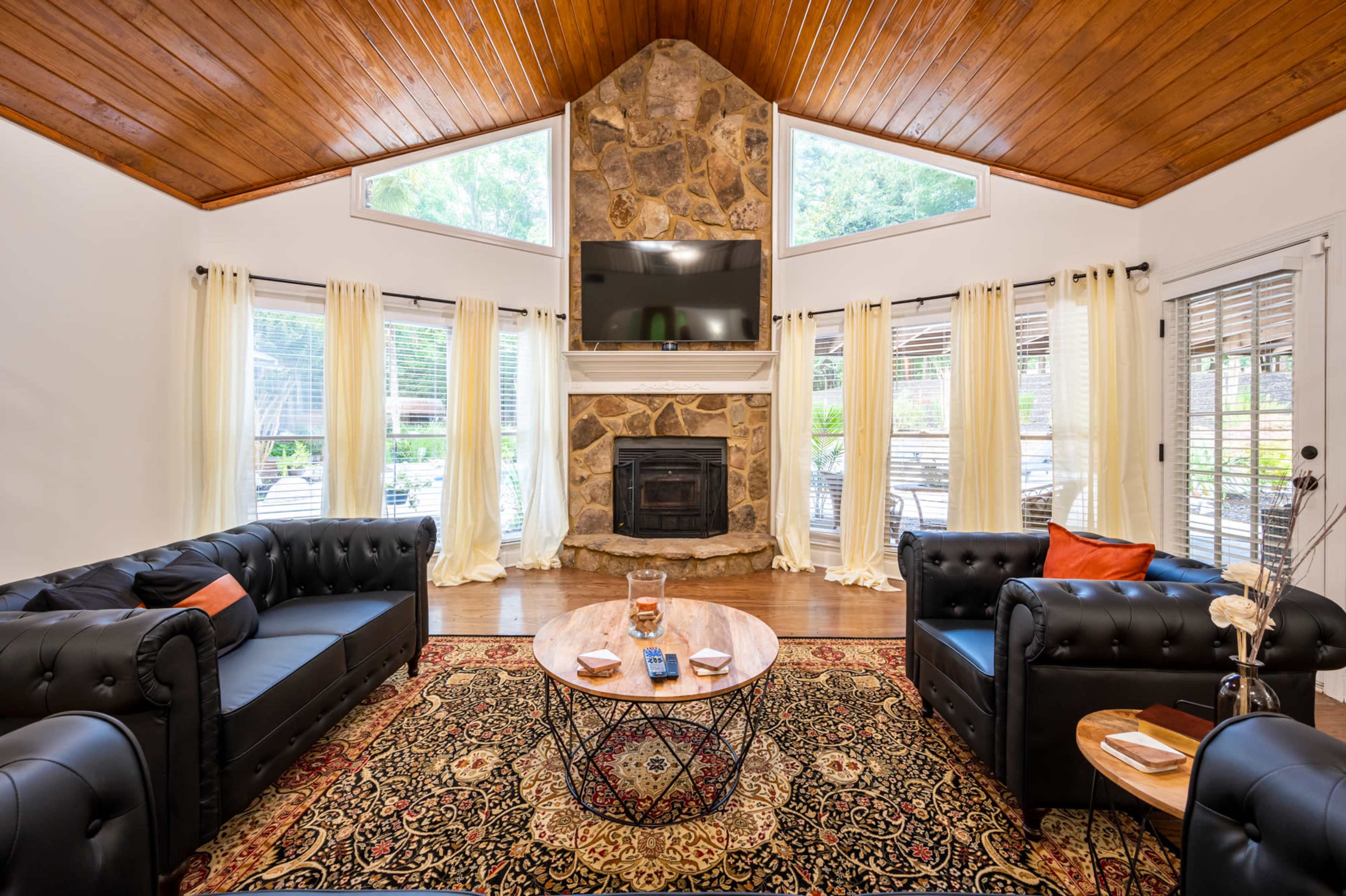 The living room features a stone fireplace, a modern television mounted above it, two black leather sofas, and a round coffee table on a patterned rug.
