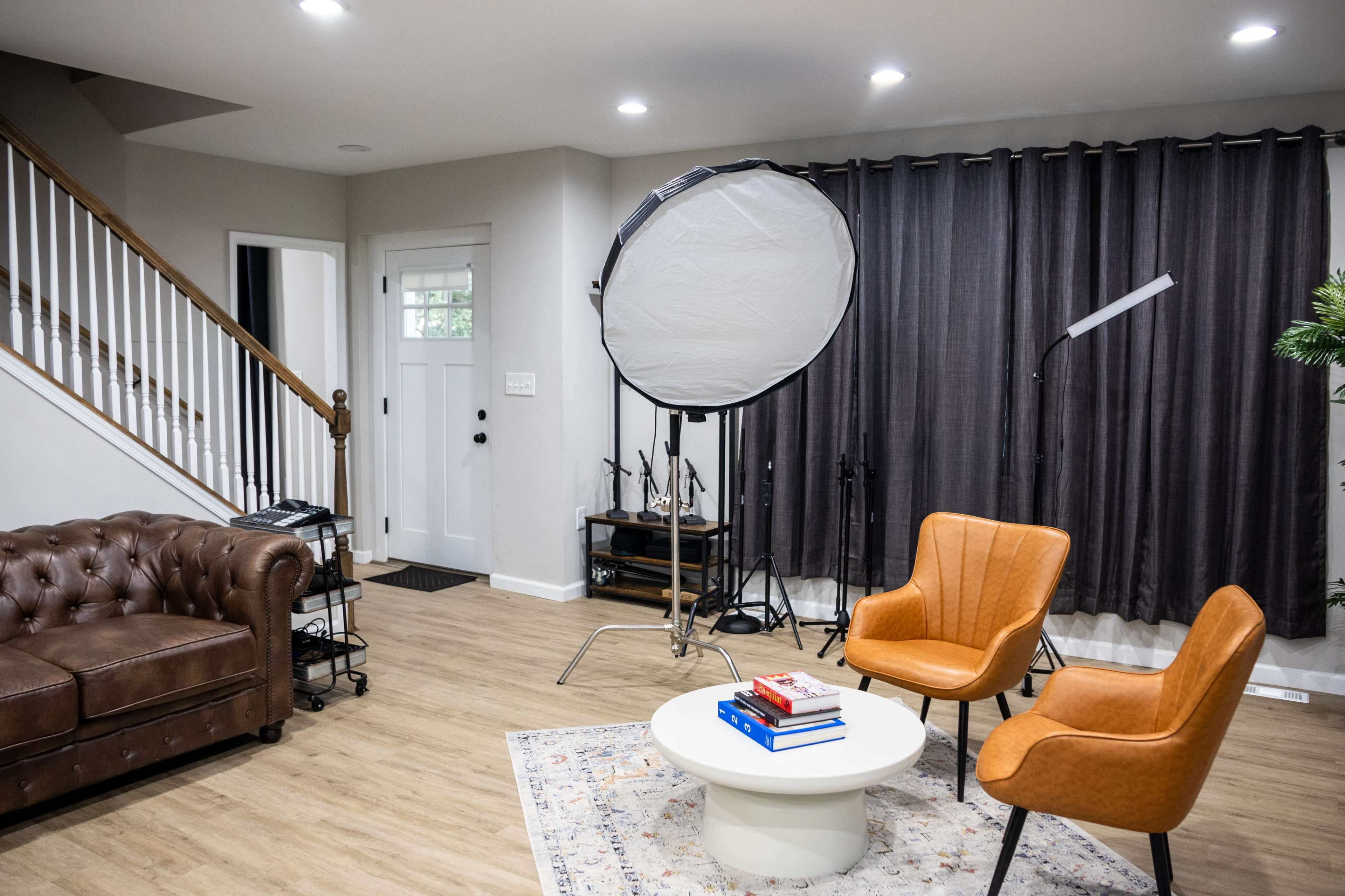 A modern living room with a brown leather couch, two orange armchairs, a round coffee table with books, and a large softbox lighting setup.