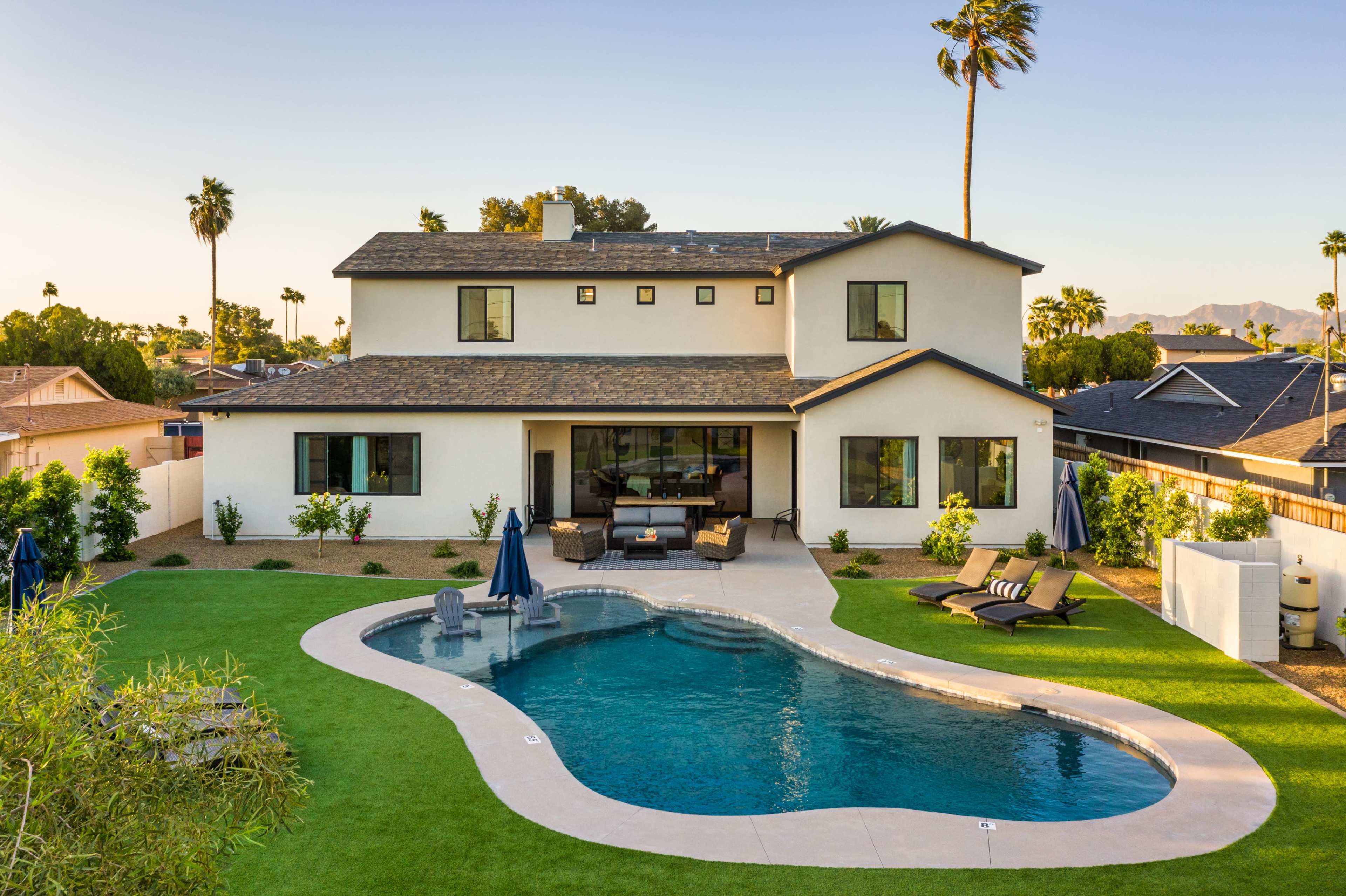 A modern two-story house features a landscaped backyard with a curved swimming pool and patio area.