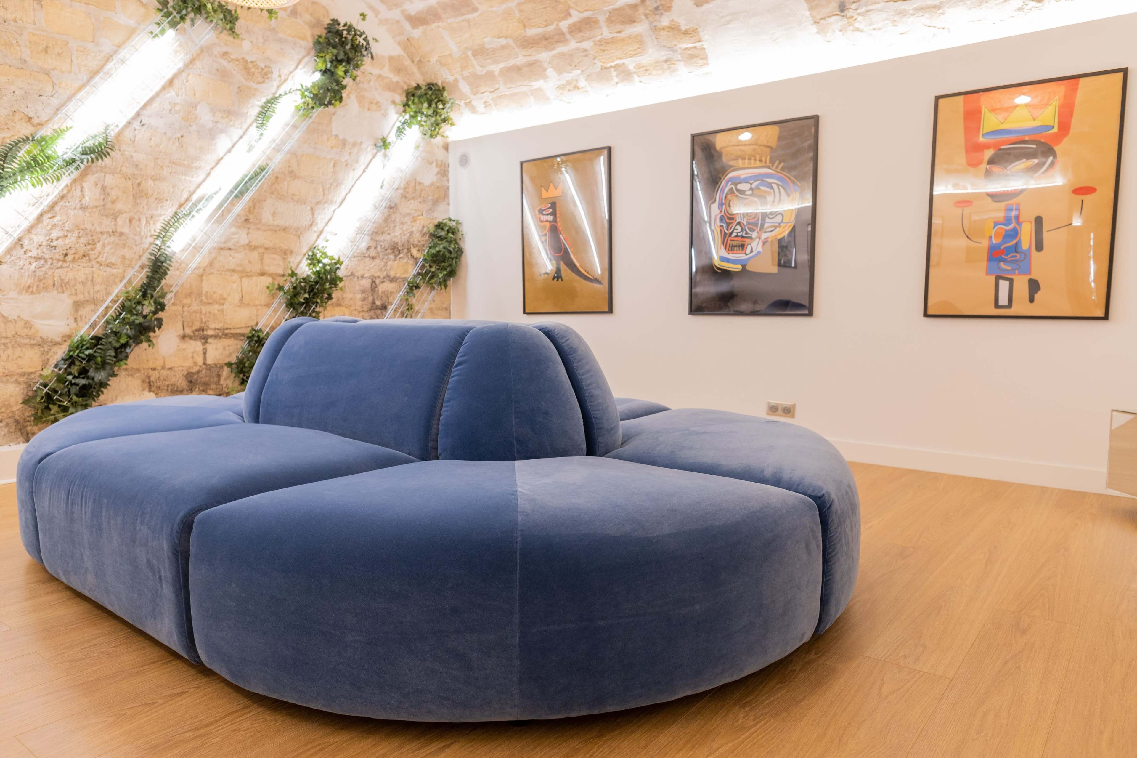 A rounded blue sofa in a minimalist room with stone walls and framed artworks hanging above it.