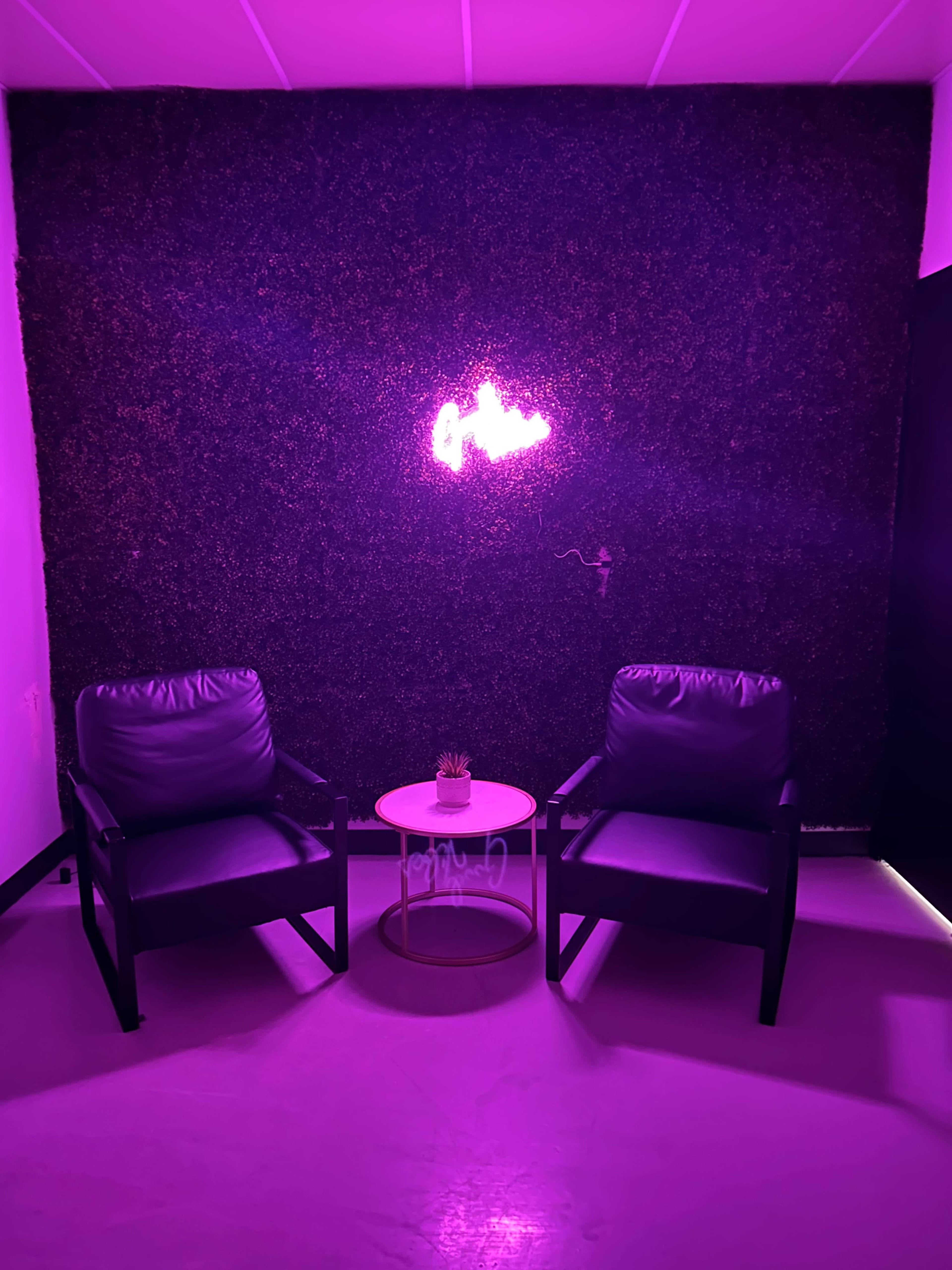 Two black chairs are positioned in front of a dark green moss wall, with a small round table in between and a neon light glowing above.
