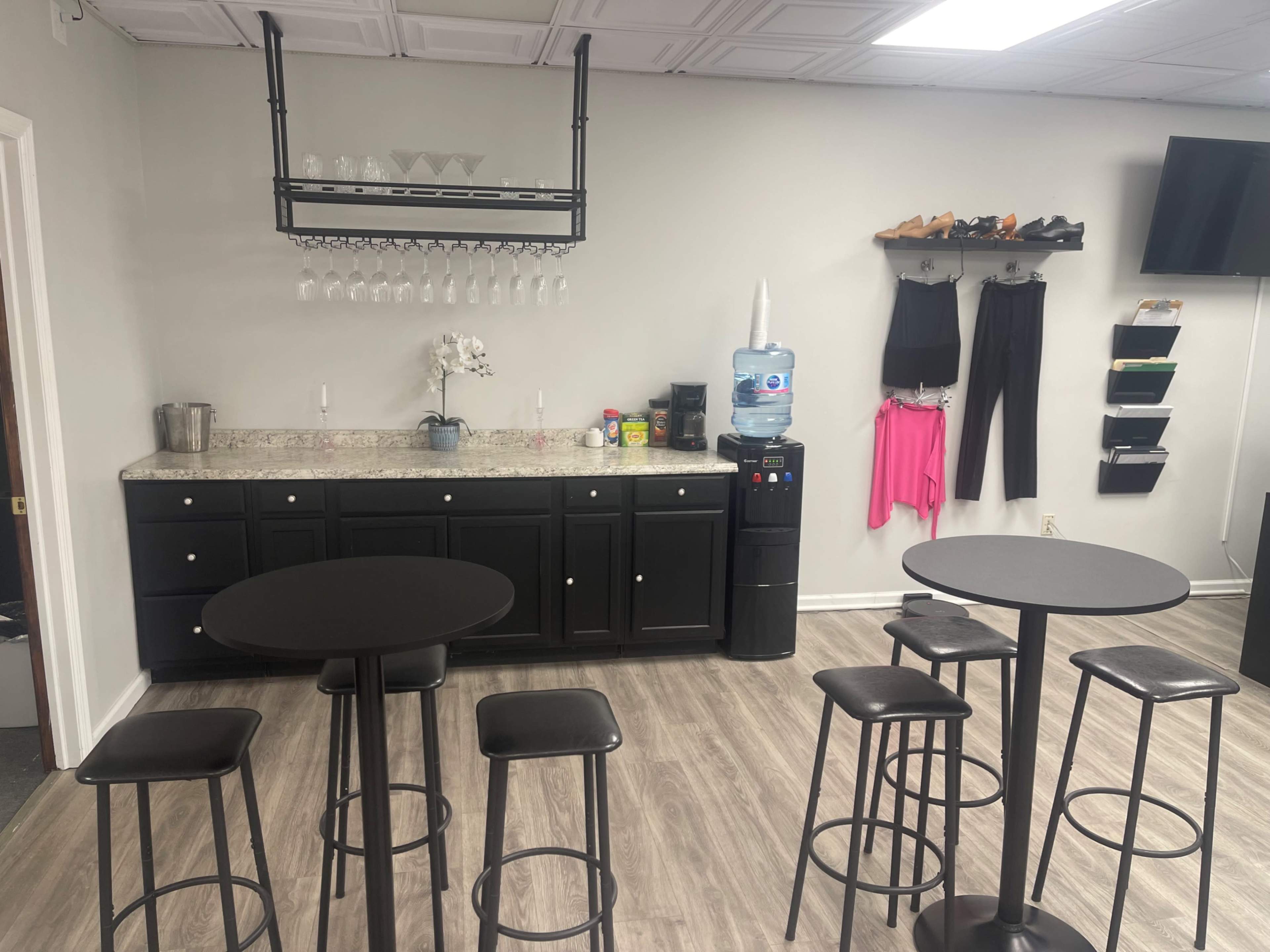 A small, modern break room featuring a black countertop with a water dispenser, glasses hanging on a rack, and several high-top tables with stools.
