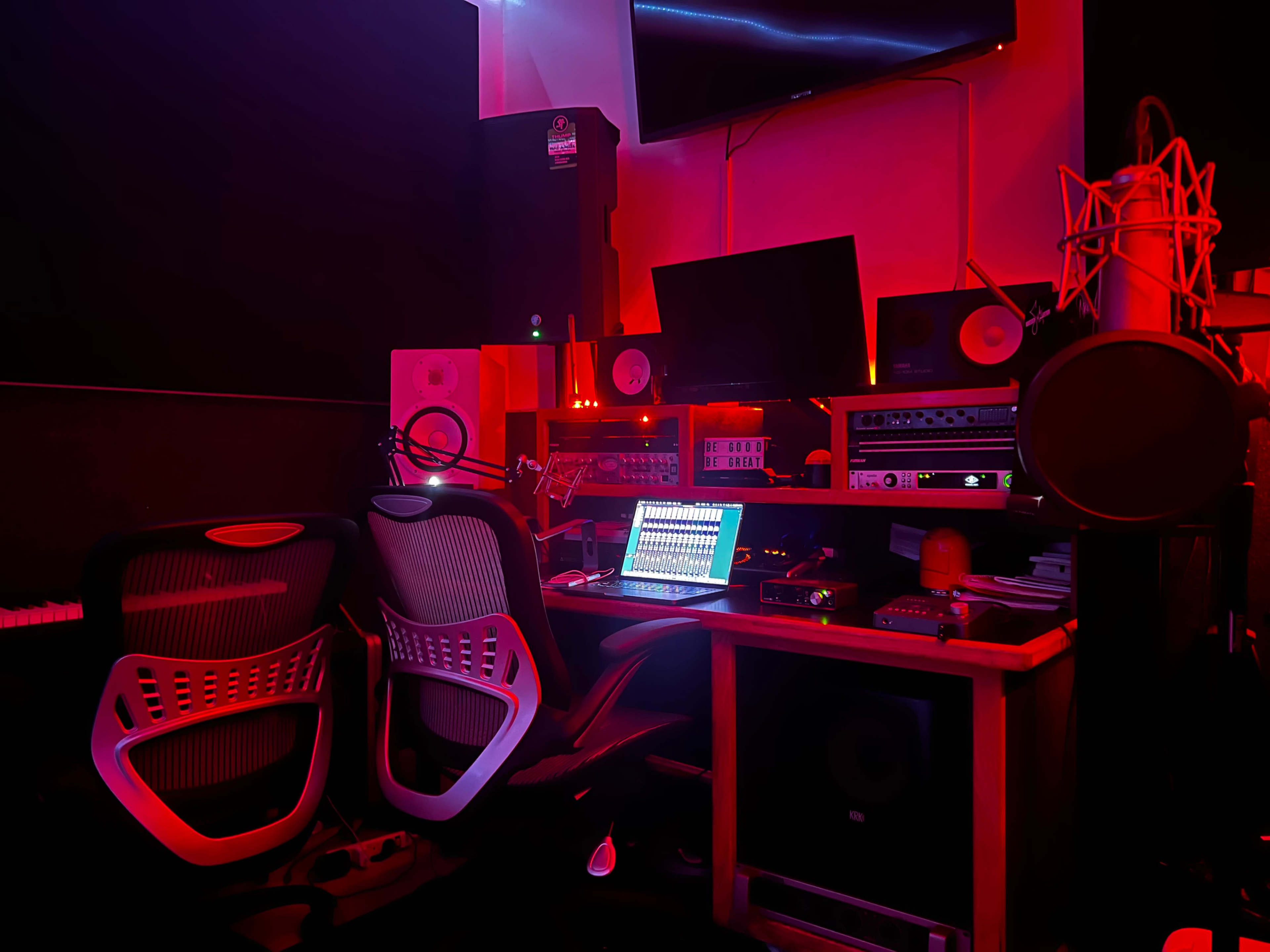 The image shows a recording studio setup with various audio equipment, a computer, and two chairs under red lighting.