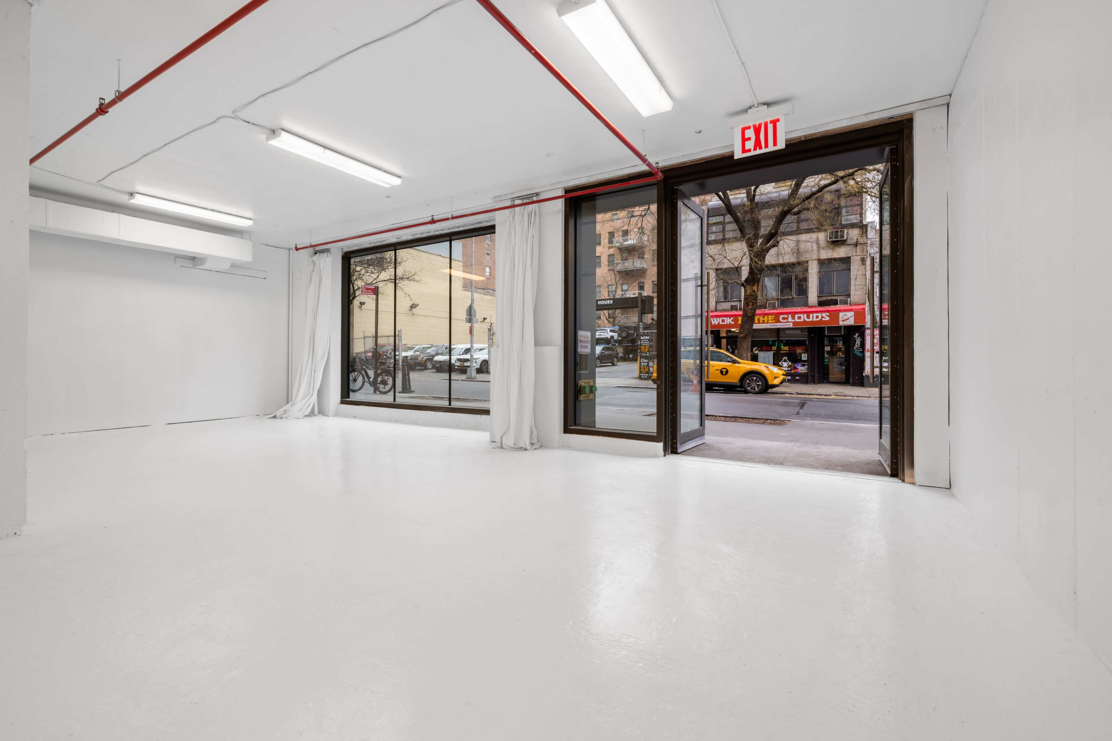 An empty interior space with white walls and a white floor, featuring large windows that open to a city street with a yellow taxi and storefronts visible outside.