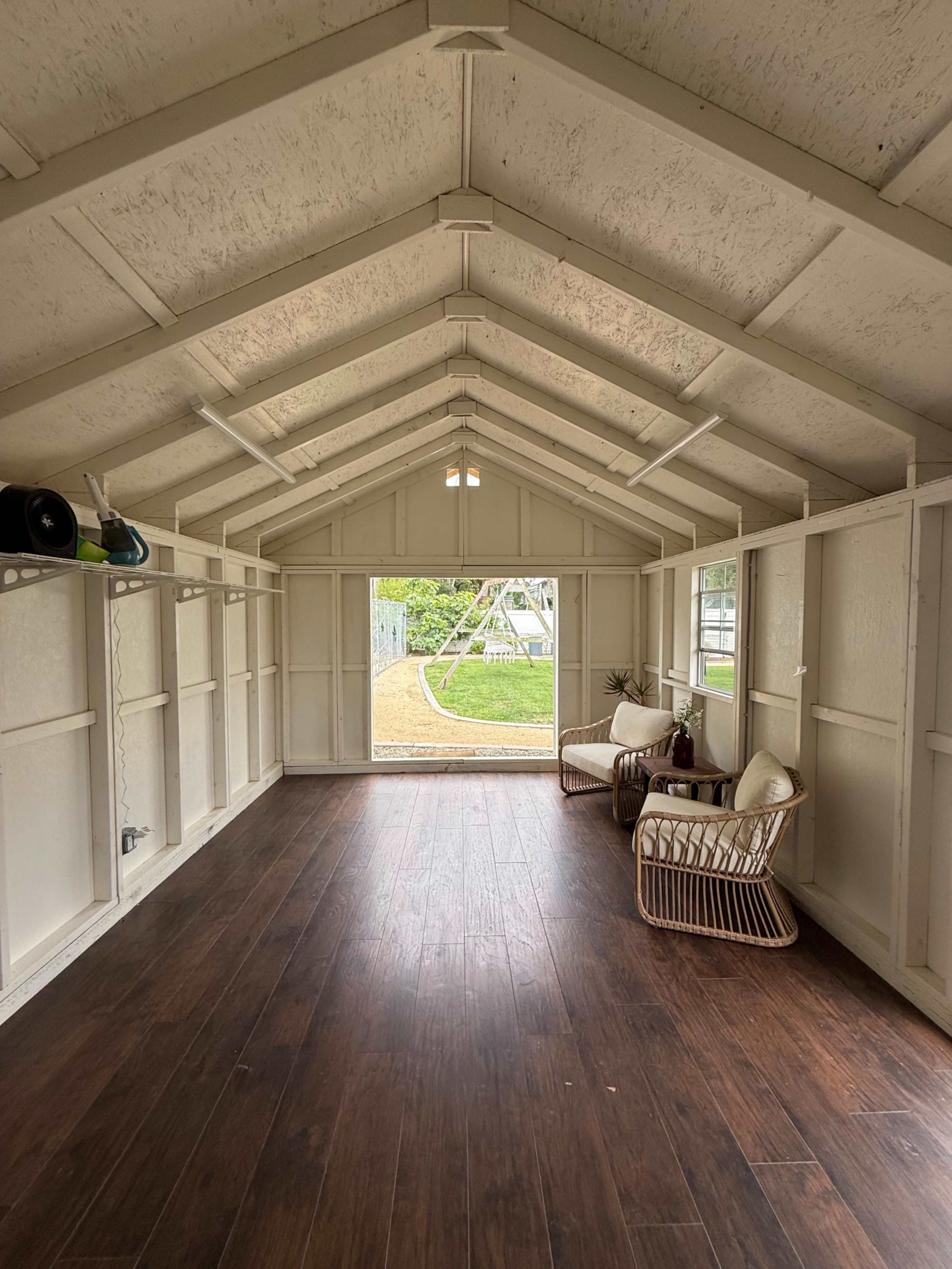 The interior of a white wooden shed features a large open space with wooden flooring, two modern chairs, and a door that opens to a yard with a swing set visible outside.