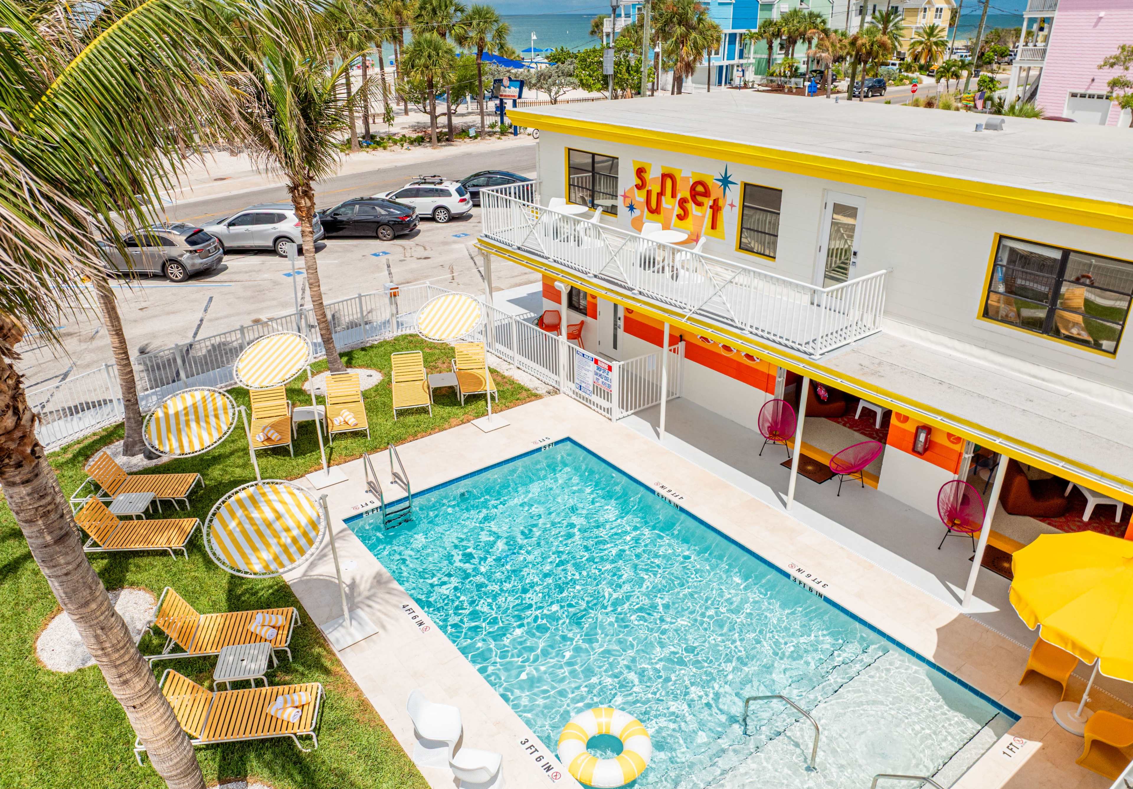 The image shows a pool area surrounded by lounge chairs and umbrellas, adjacent to a brightly colored building with a sign that reads "Sunset."