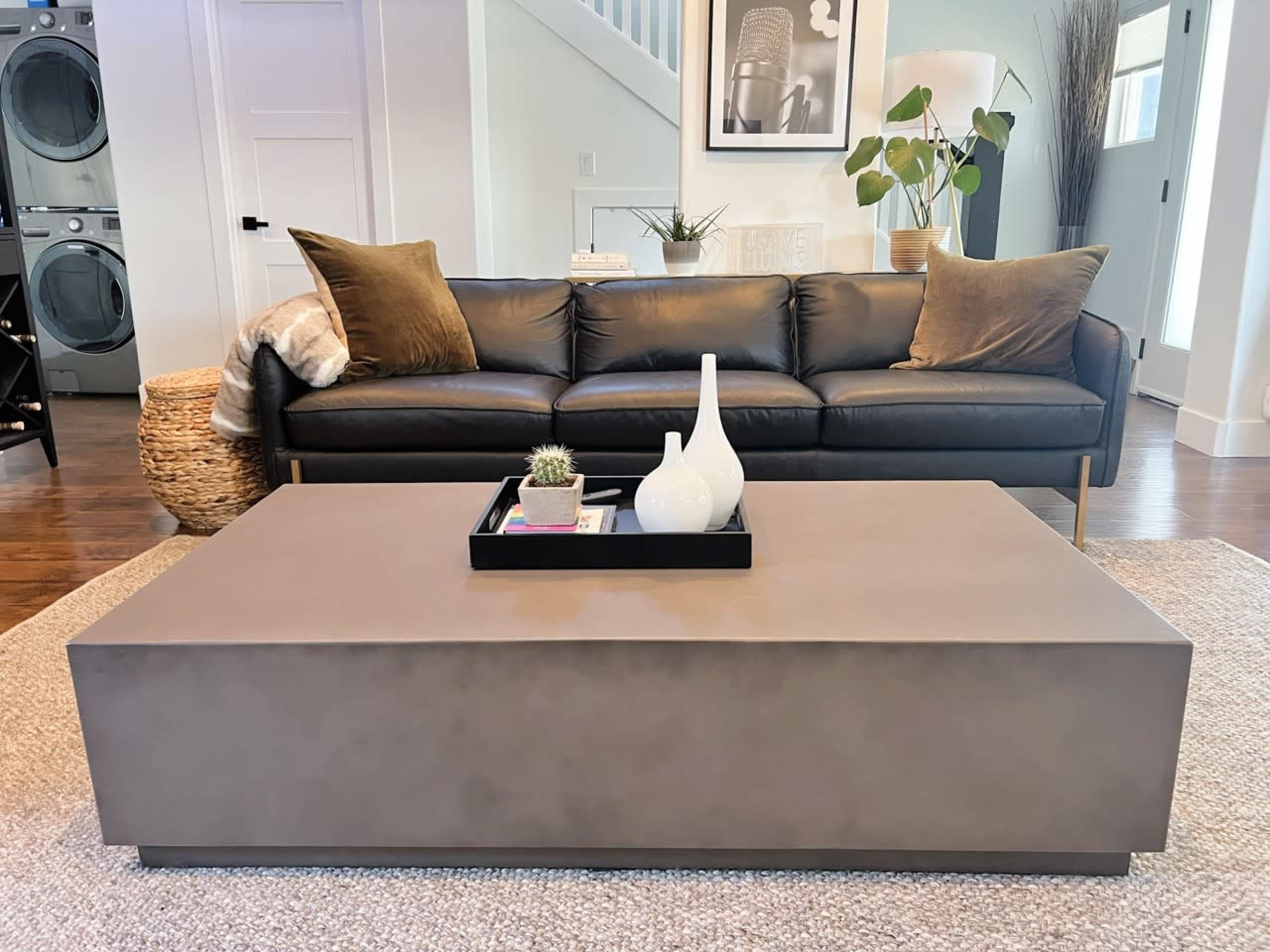 A black leather sofa with decorative pillows sits in front of a rectangular gray coffee table on a light-colored rug in a modern living room.