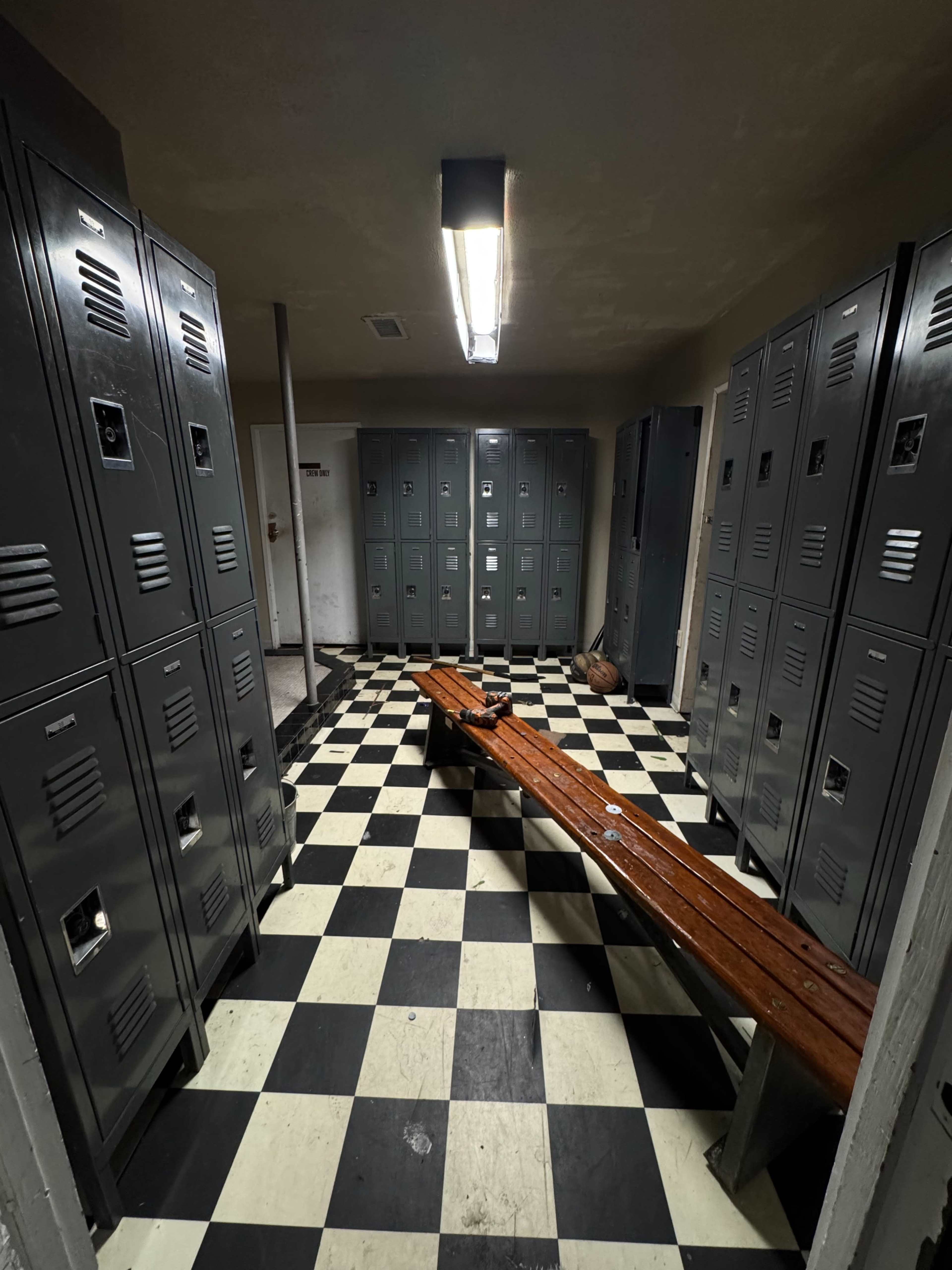 The image shows a dimly lit locker room featuring gray metal lockers lining the walls and a wooden bench in the center on a black and white checkered floor.