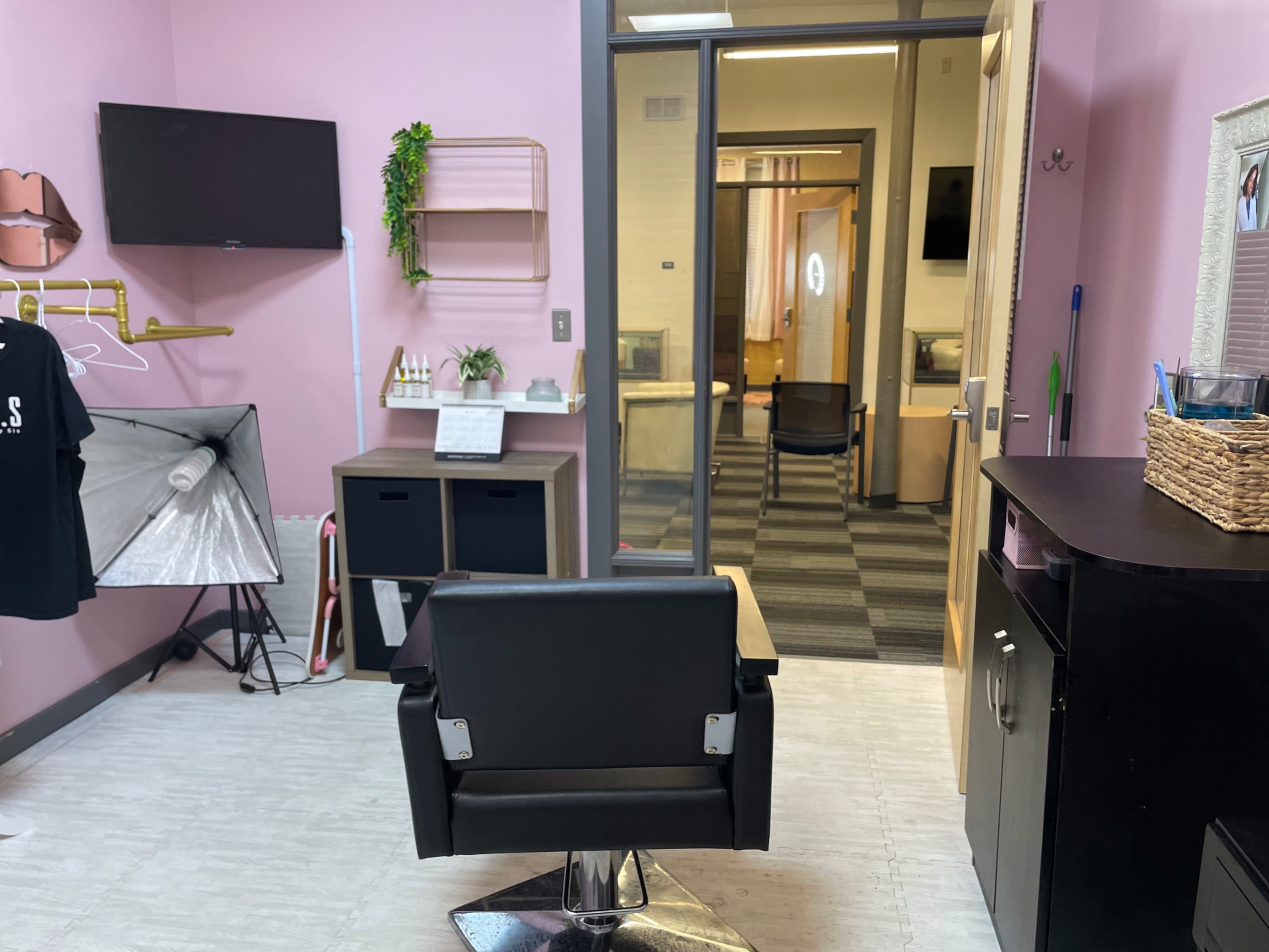 A brightly colored salon room with a black styling chair, a mirror, and storage units, leading into additional workspaces through an open door.