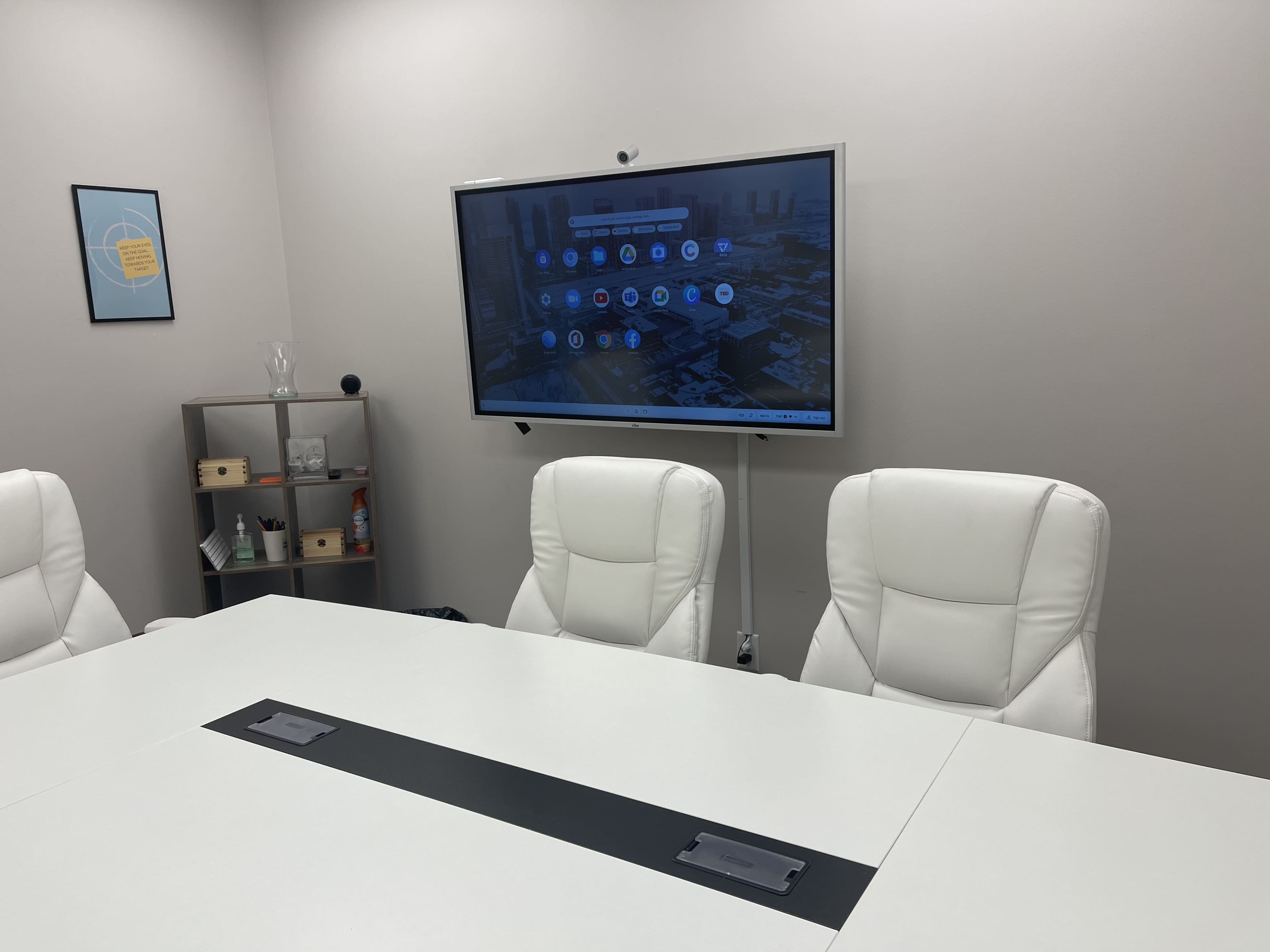 A modern conference room with a large screen mounted on the wall and a white table surrounded by three white chairs.