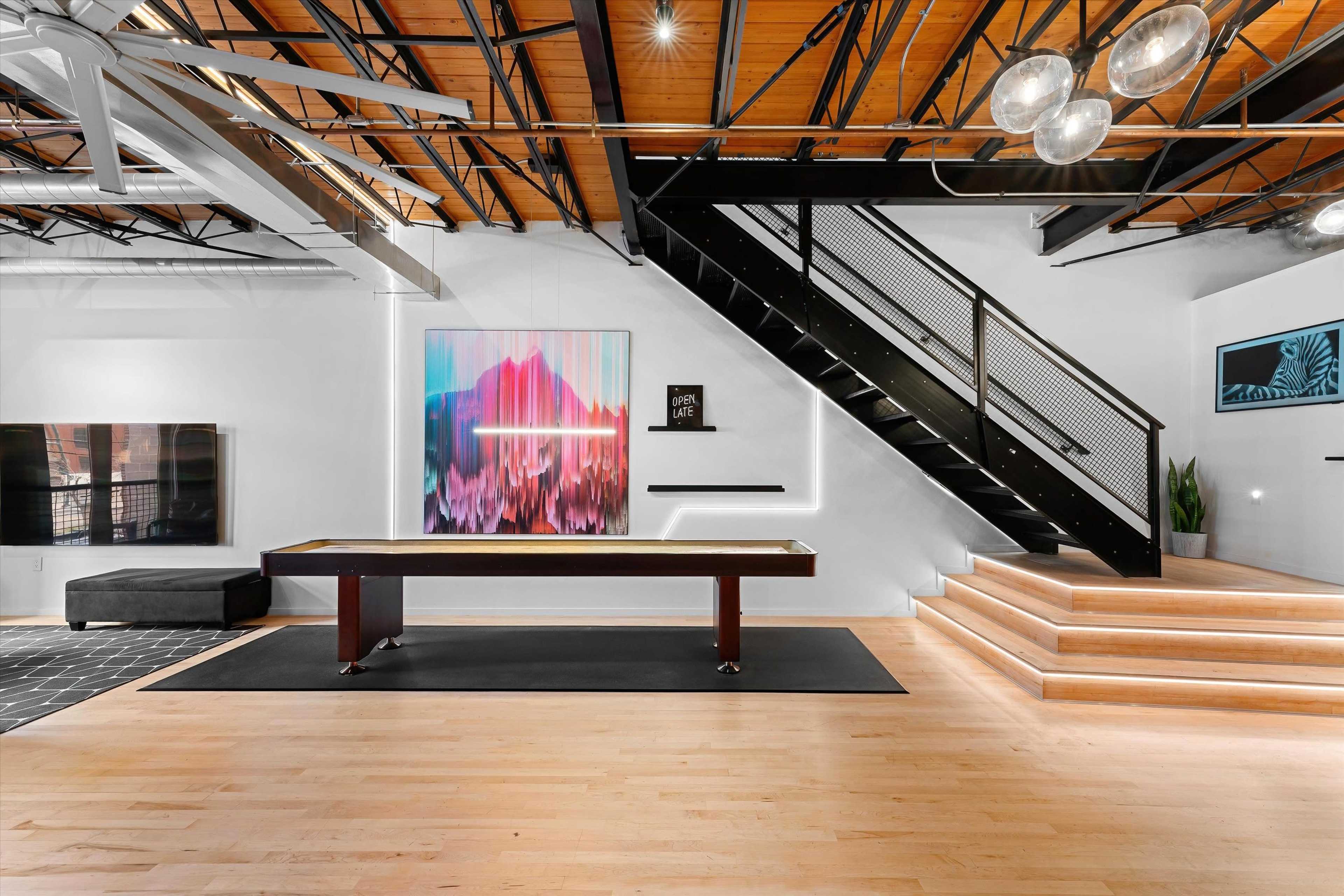The image shows a modern interior space featuring a pool table, a staircase with black railings, and colorful artwork on the wall.