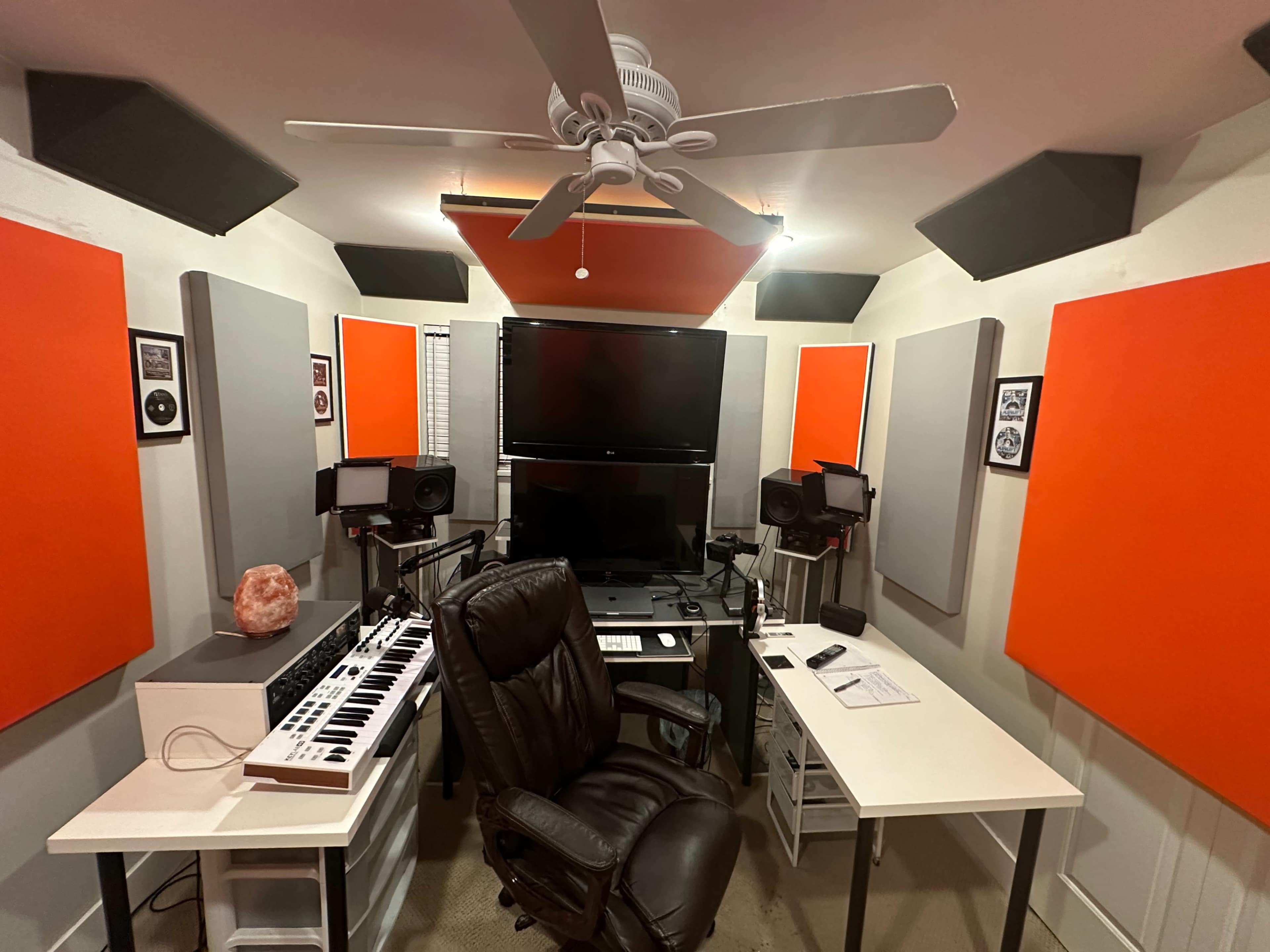 A recording studio with a black chair, computer monitors, a MIDI keyboard, and orange acoustic panels on the walls.