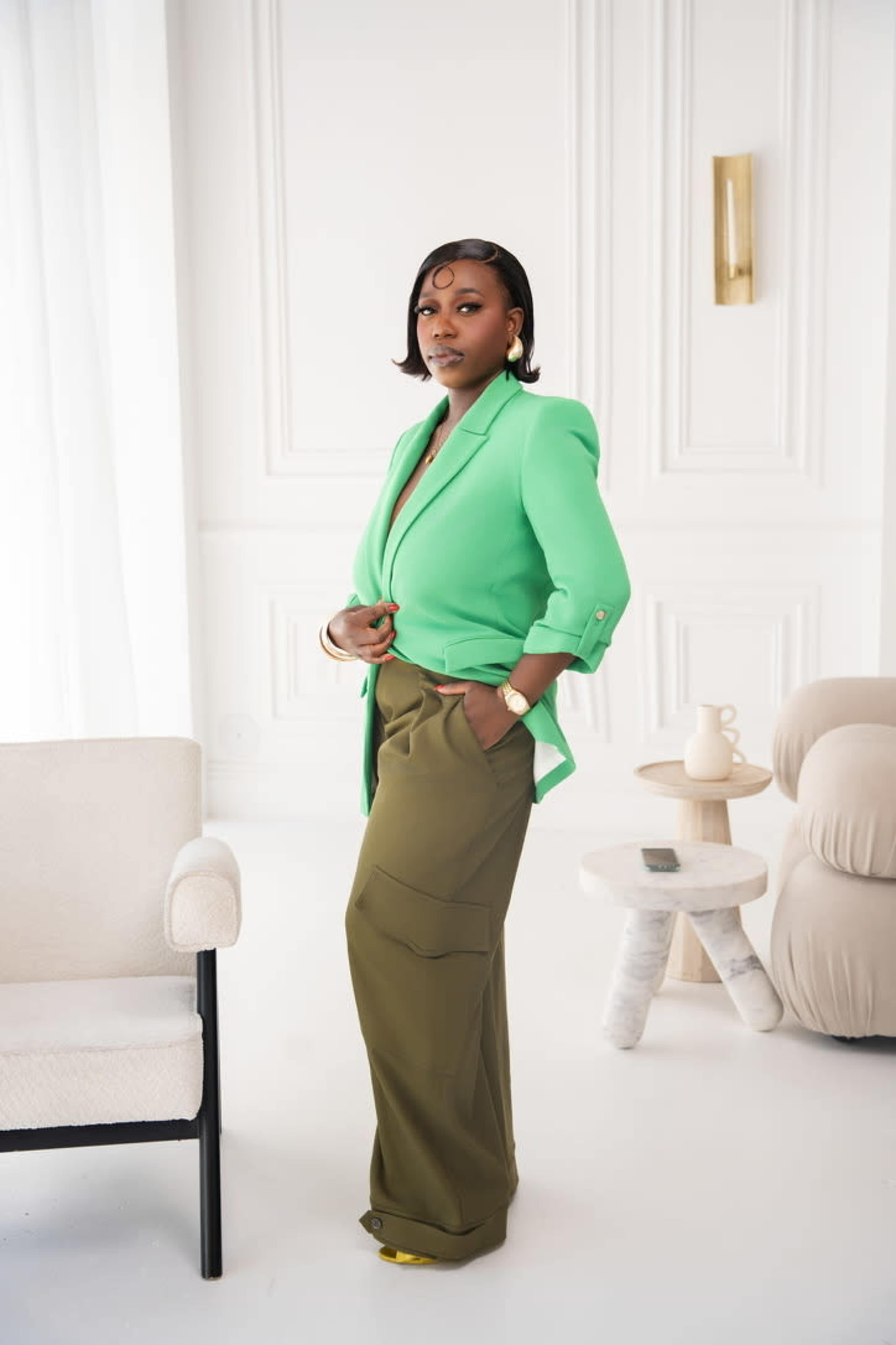 A woman in a bright green blazer and olive cargo pants stands in a modern, minimally furnished room.