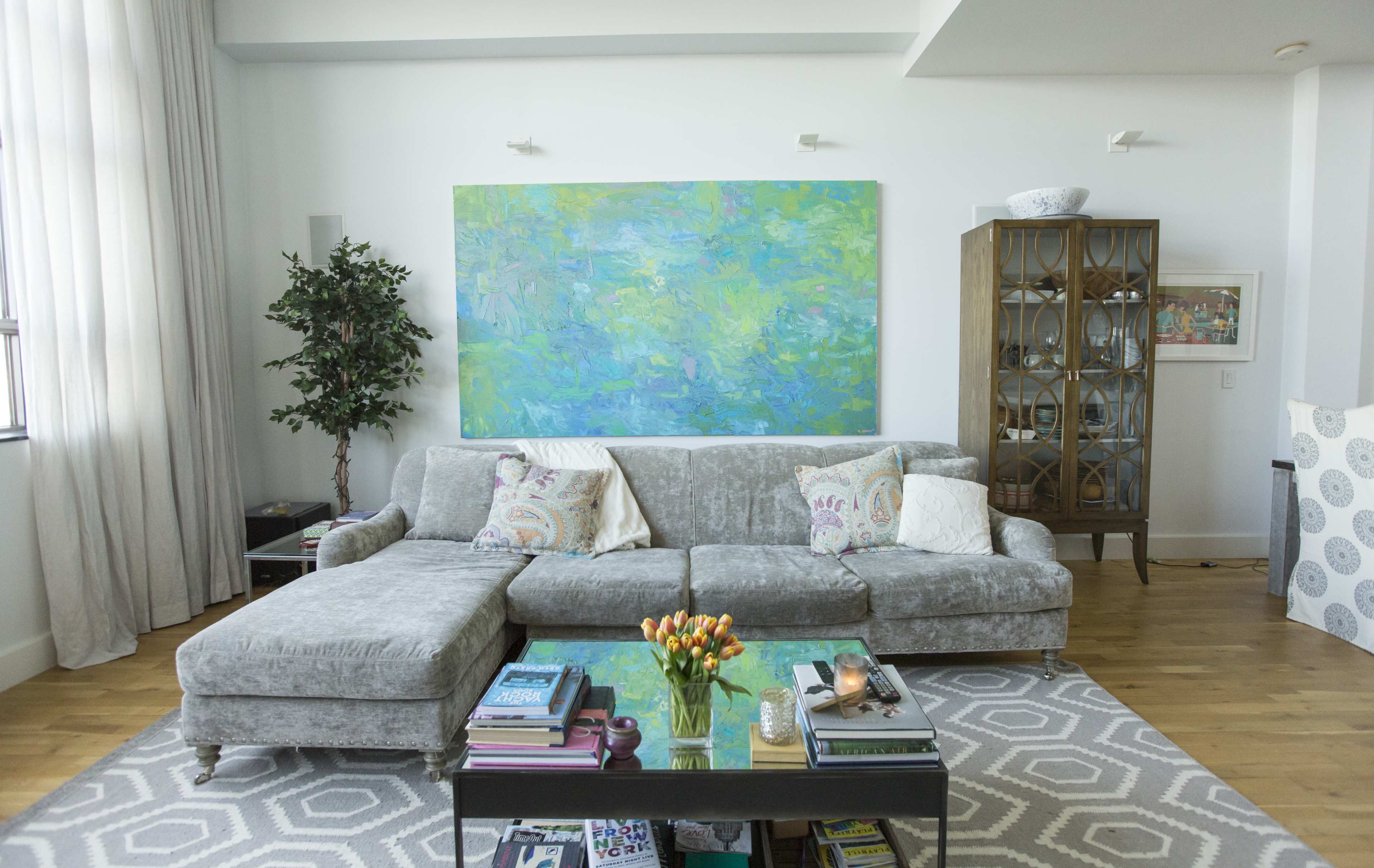 A modern living room features a gray sectional sofa, a glass coffee table with books and decor, and a large abstract painting on the wall.