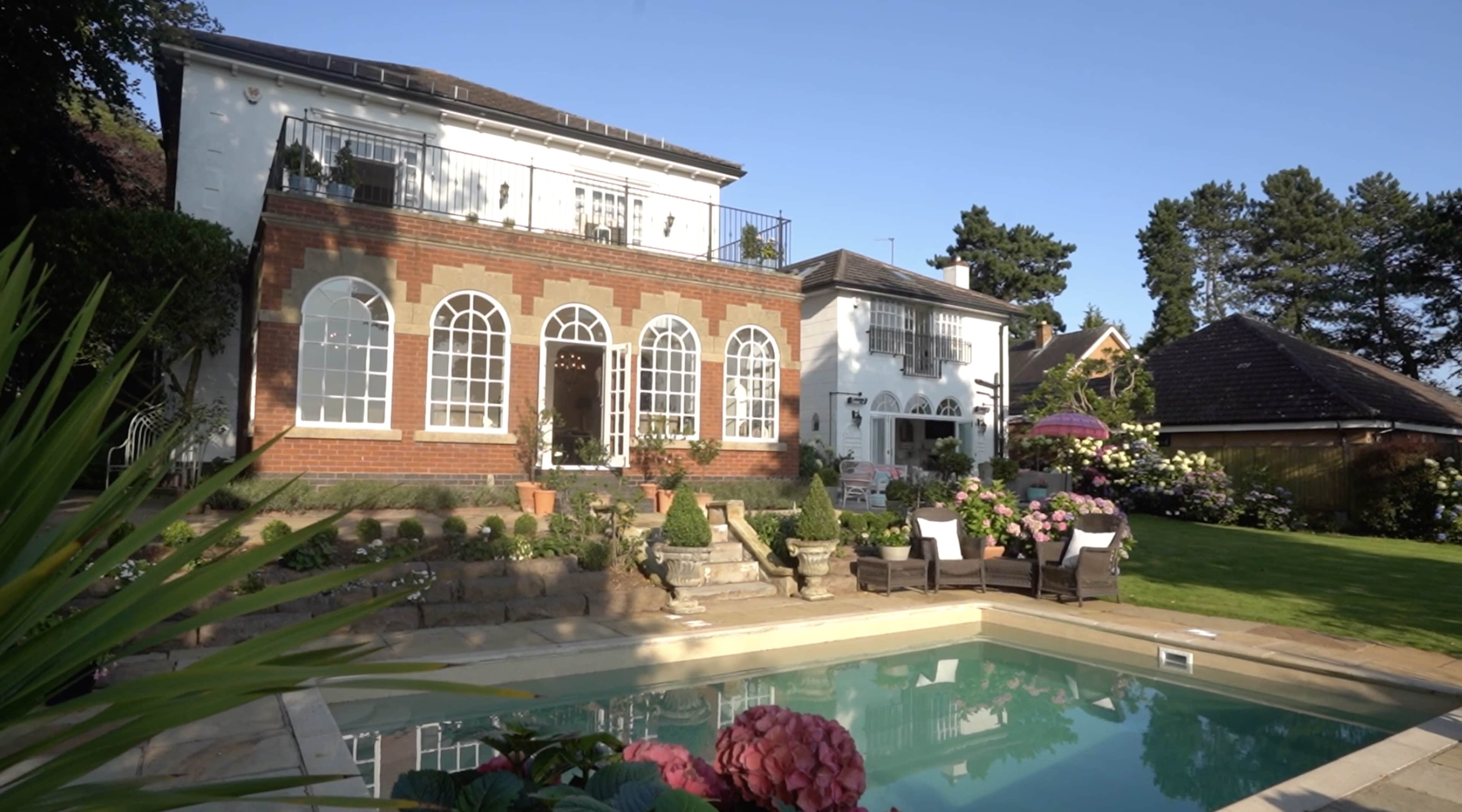 A modern house with large arched windows overlooks a swimming pool surrounded by meticulously landscaped gardens.