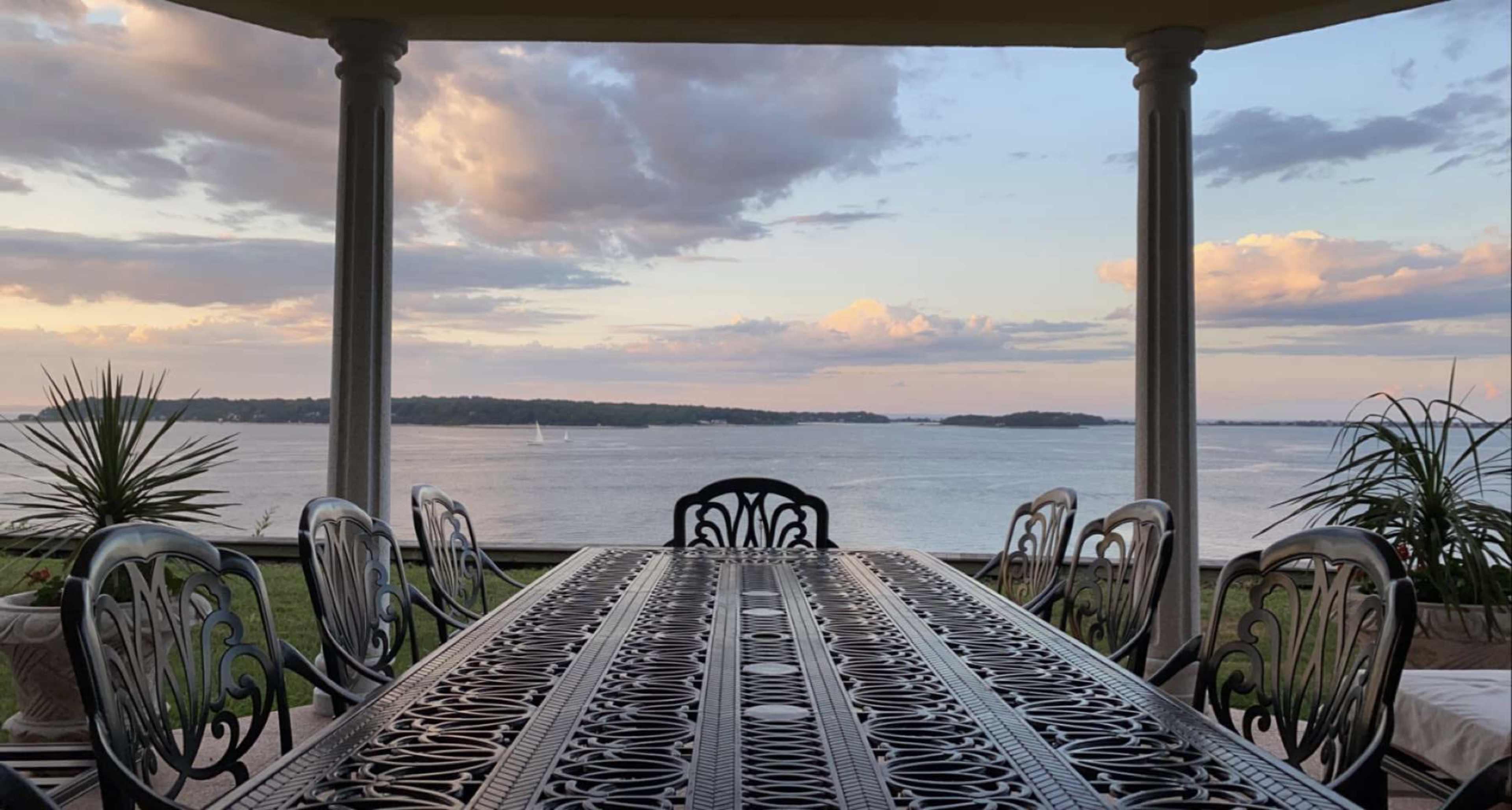 A waterfront patio features a sculpted metal table surrounded by chairs, with a view of the water and clouds above.