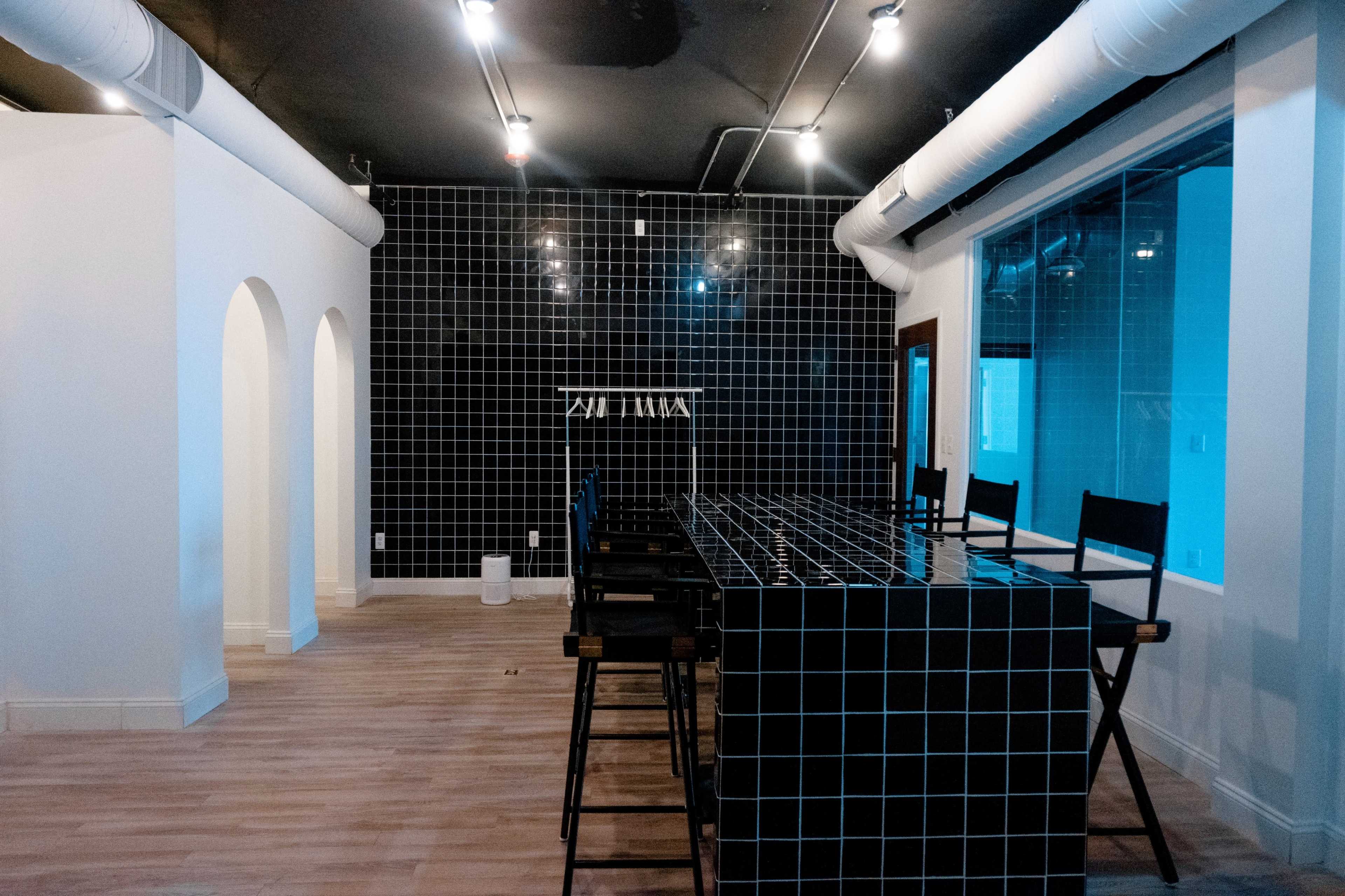 An empty room with a black tiled wall, a long black tiled bar, and several high stools arranged in front of it.