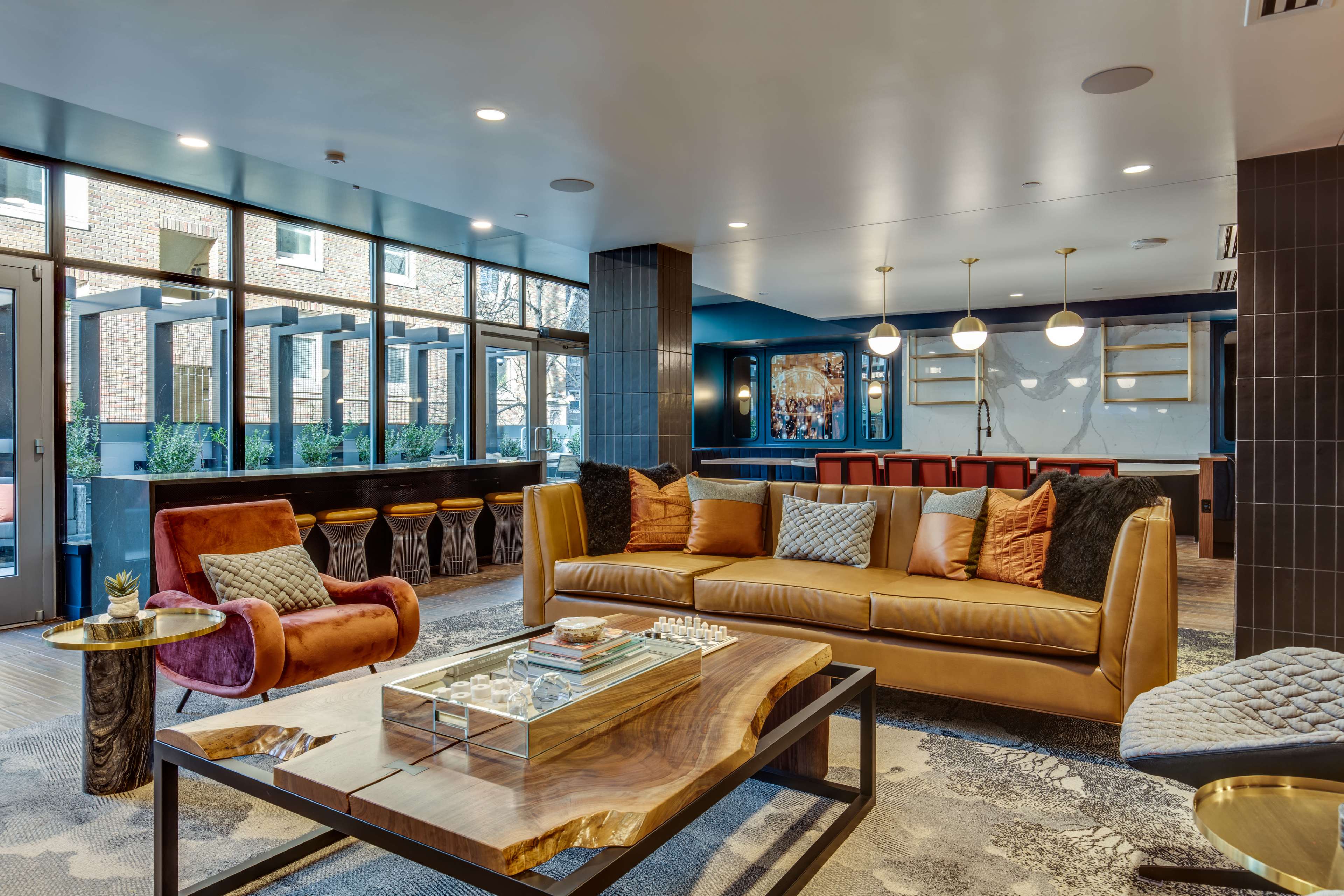 A modern lounge area featuring a brown leather sofa, a red accent chair, a wooden coffee table, and large windows letting in natural light.